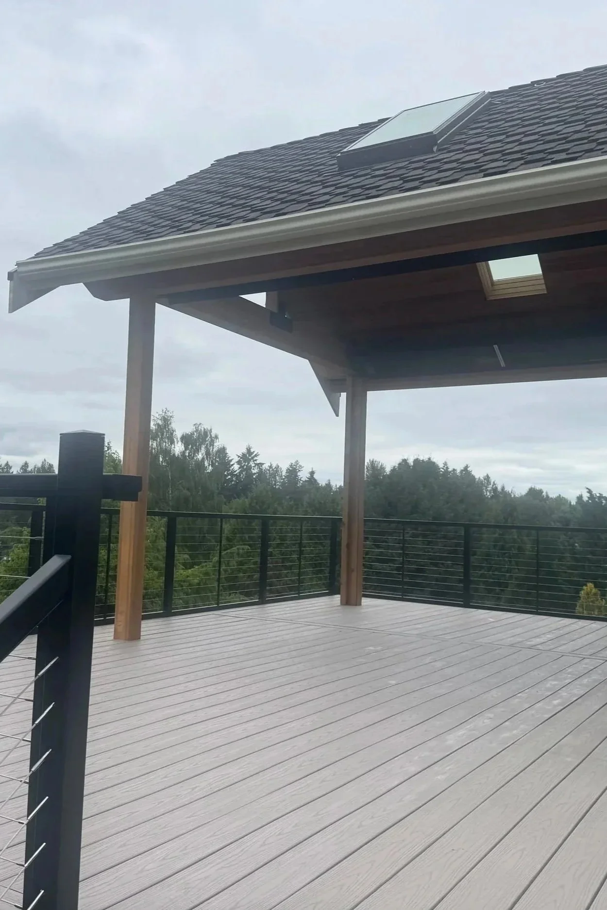 A spacious outdoor deck with wooden flooring, surrounded by a black metal railing, featuring a partial view of a house roof with a skylight and a cloudy sky in the background.