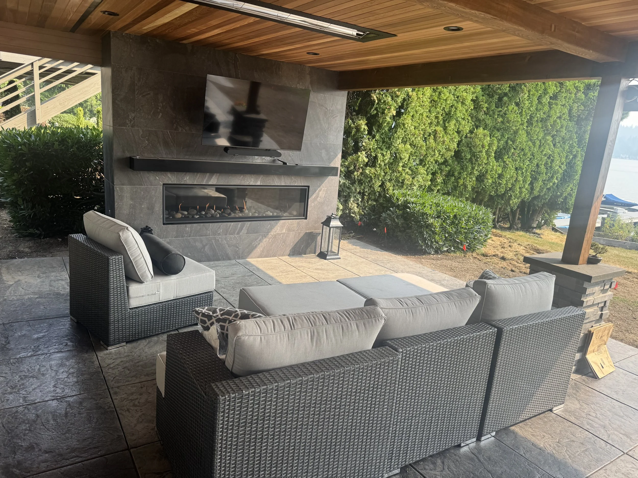 Outdoor patio area with a wall-mounted flat-screen TV, a modern fireplace beneath it, and cozy seating including a black wicker chair with a beige cushion and a matching sofa. There are green trees and a view of the water in the background.