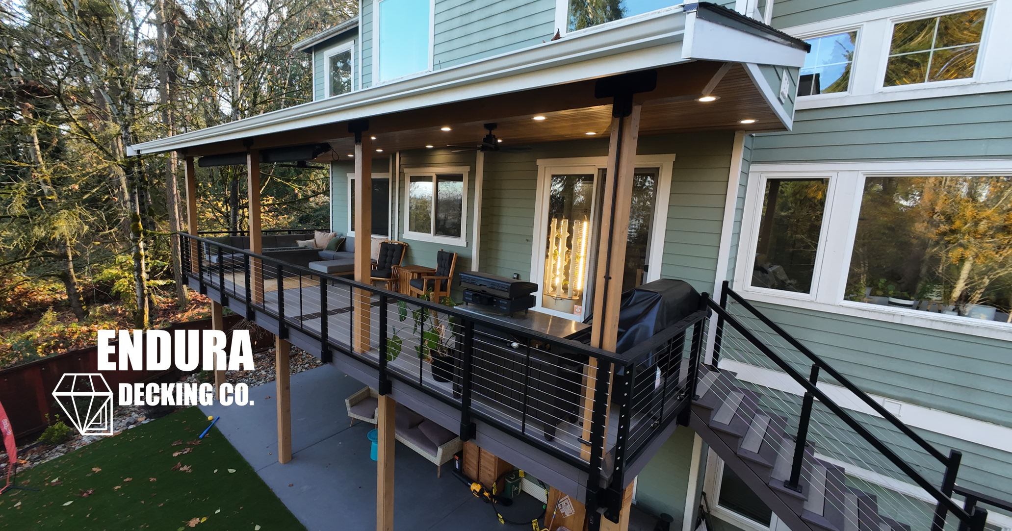 A two-story house with a spacious elevated outdoor deck, featuring outdoor furniture, a barbecue grill, and string lights, surrounded by trees.