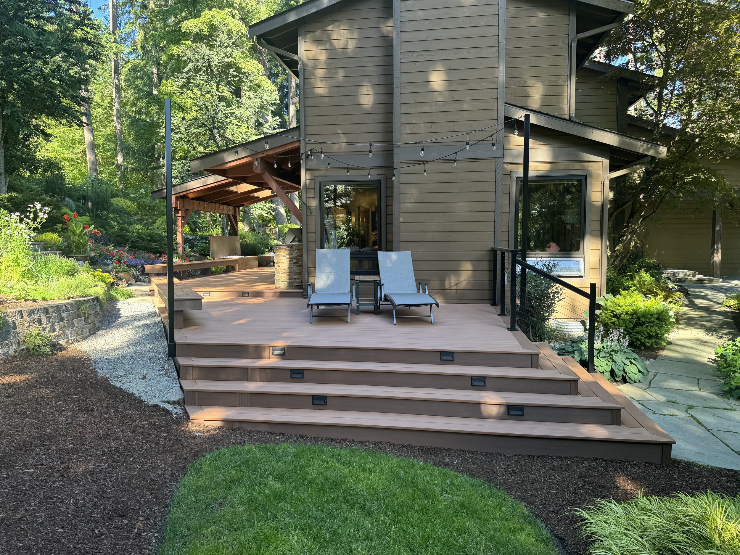 A two-story house with exterior wood siding, surrounded by lush green trees and landscaping. In front of the house, there is a wooden deck with outdoor furniture and string lights hanging above. A gravel pathway borders the deck, leading to a garden 
