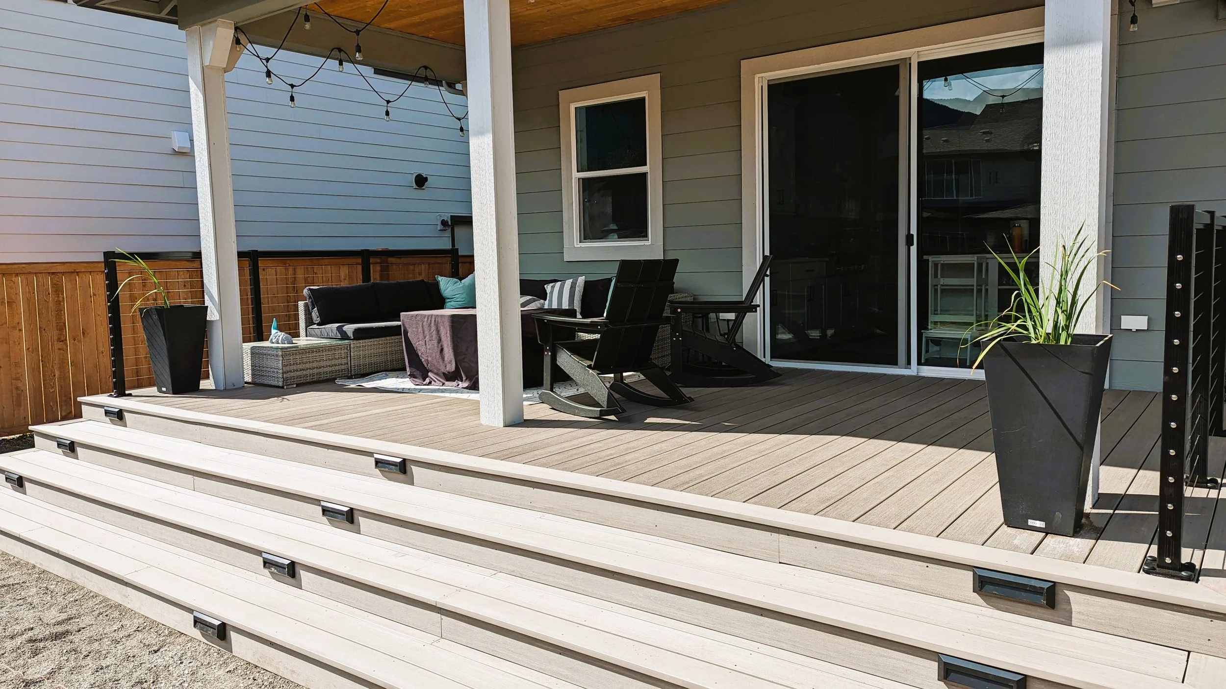 A backyard patio with light-colored wooden steps leading up to a deck area furnished with black chairs, a wicker sofa with cushions, and potted plants. There are string lights hanging from the ceiling and sliding glass doors leading inside the house.