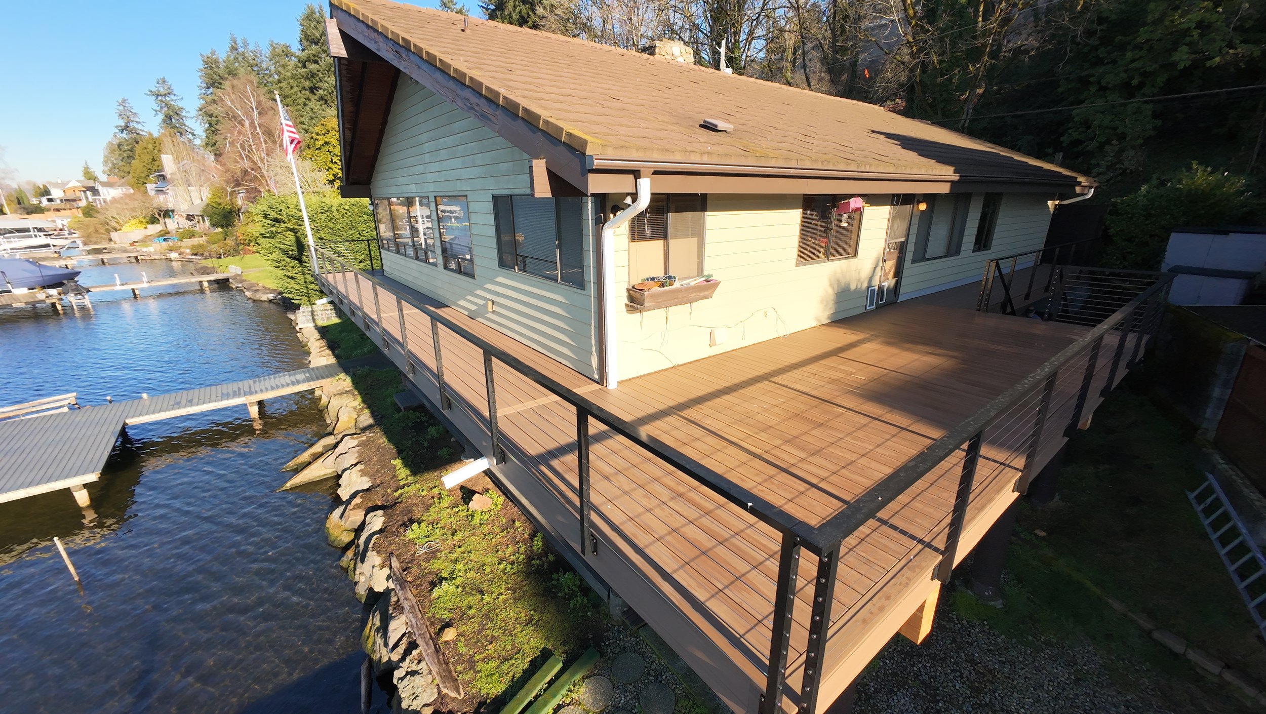 A house on stilts with a large wooden deck overlooking a body of water, surrounded by trees and outdoor scenery.