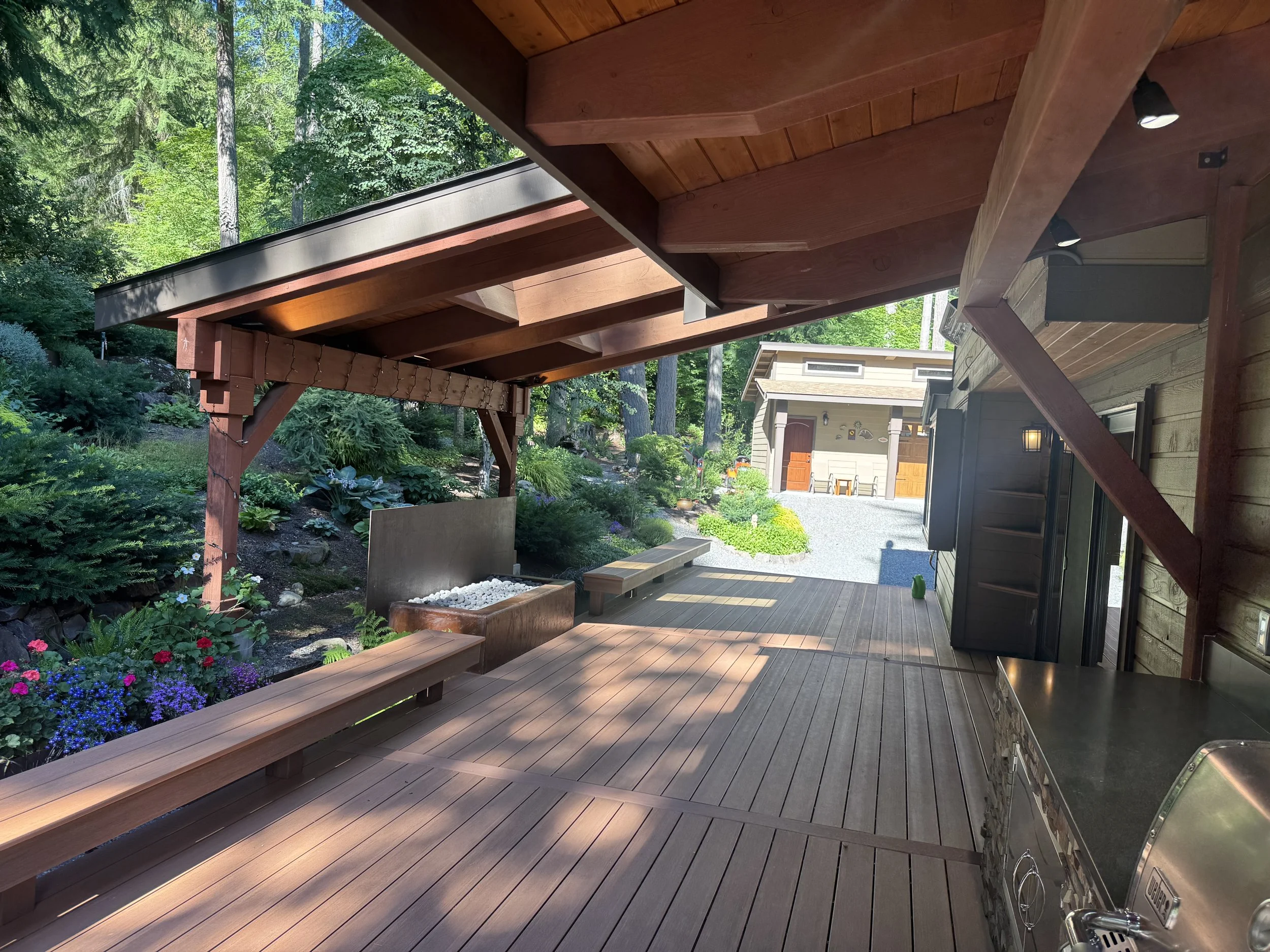 A wooden deck attached to a house, with a garden and trees in the background. There is an outdoor grill, a bench, and a small shed visible.