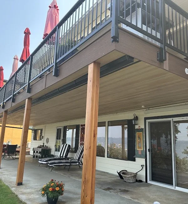 View of a deck with black metal railings and pink umbrellas on top, supported by wooden posts, overlooking outdoor patio furniture, including two striped lounge chairs, with a house exterior featuring large windows and sliding door, and a potted plan