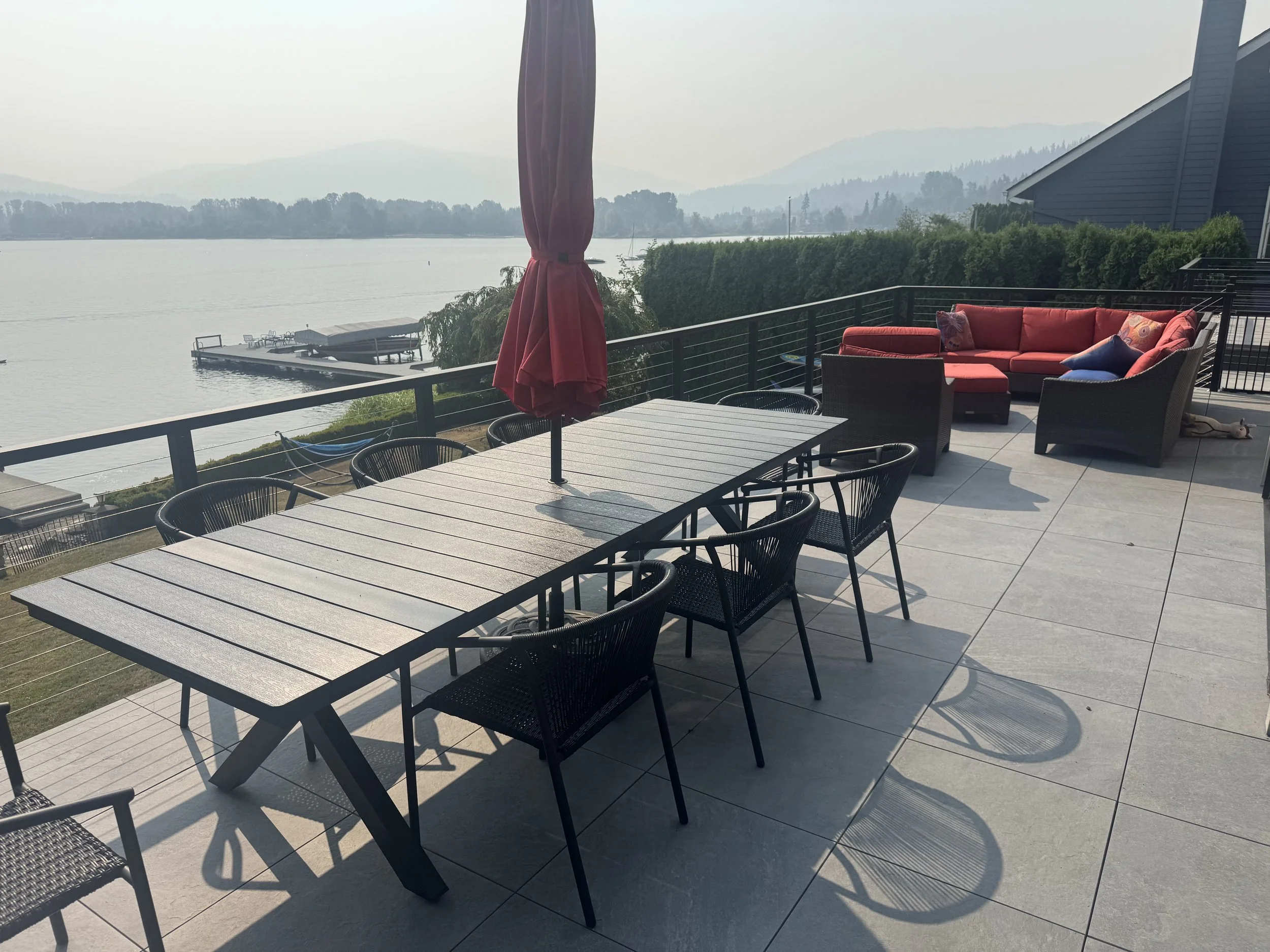 A lakeside patio with a dining table, chairs, a red umbrella, a cushioned outdoor sofa, and a scenic view of water, trees, and distant mountains.