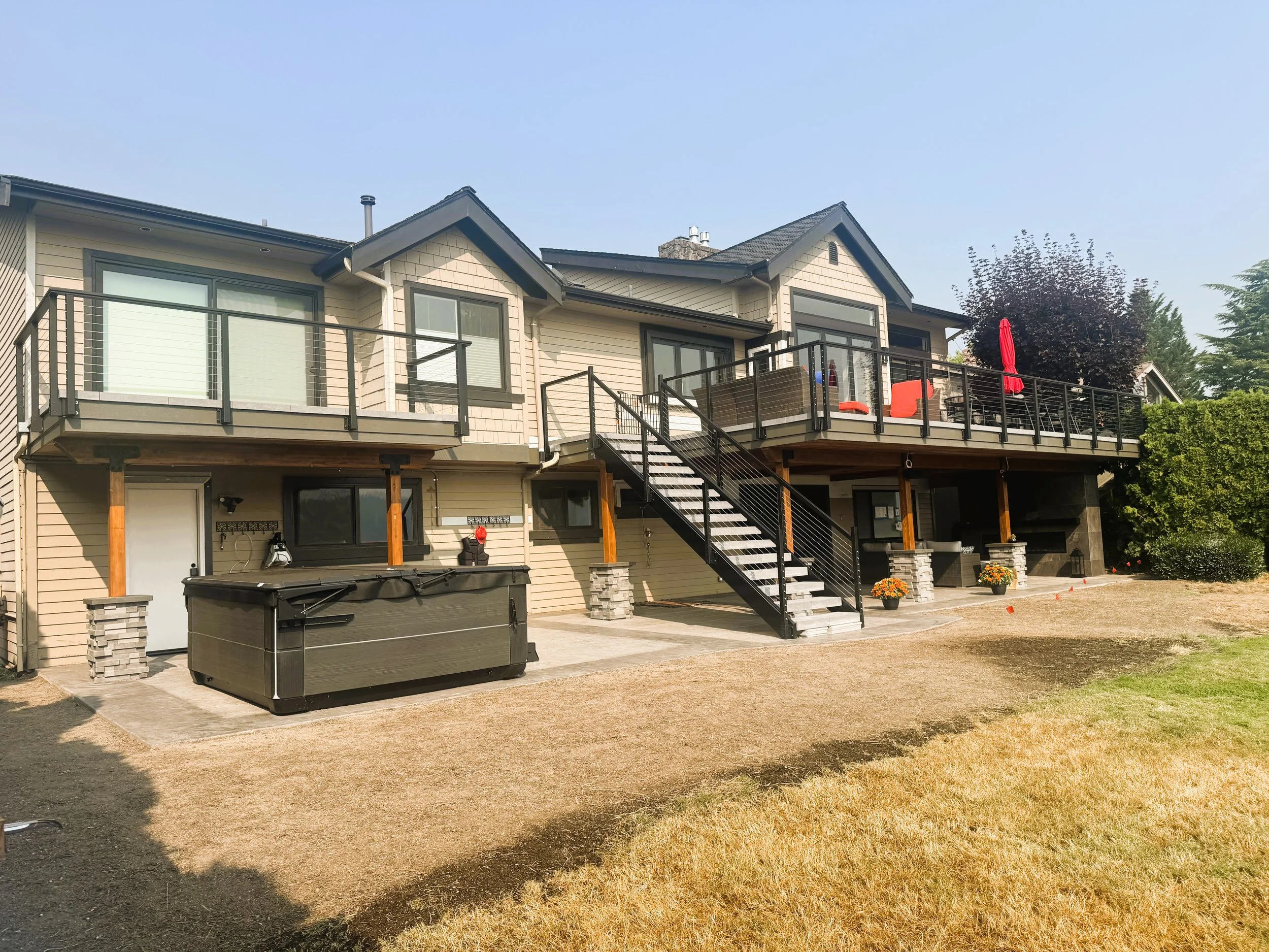 Rear view of a two-story house with a balcony, stairs, and a yard with dry grass and some potted plants.