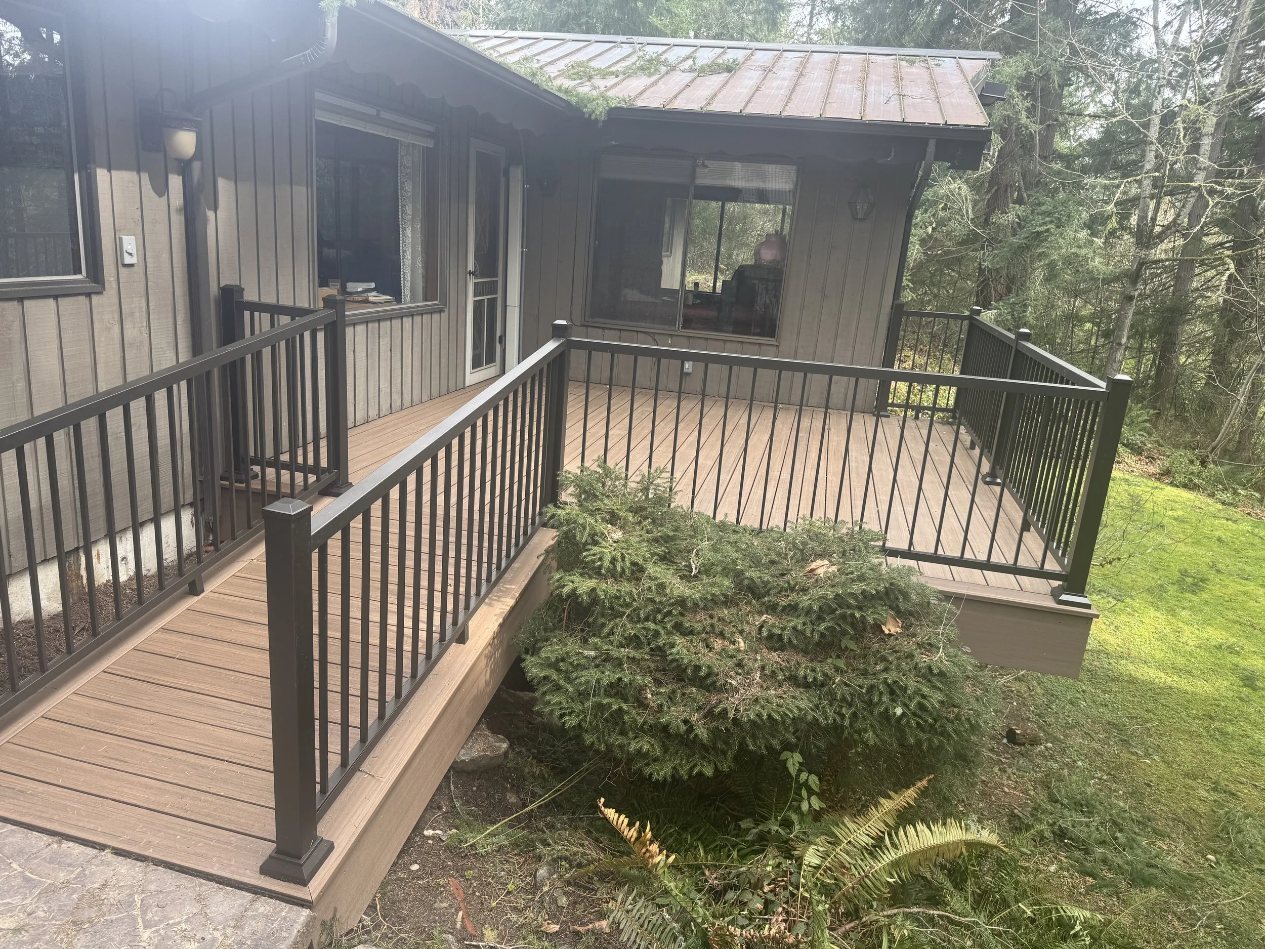 Wooden deck with black metal railing attached to a house, surrounded by lush green trees and plants.