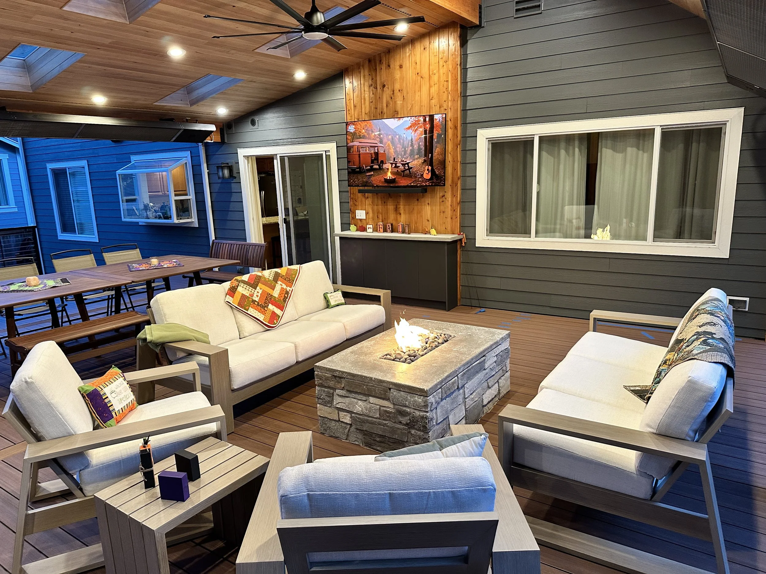 Outdoor covered patio with wooden ceiling, ceiling fan, recessed lighting, gray wooden walls, large window, sliding glass door, and a TV mounted on the wall. Furnished with cream-colored seating, a wooden dining table, and a fire pit table in the cen
