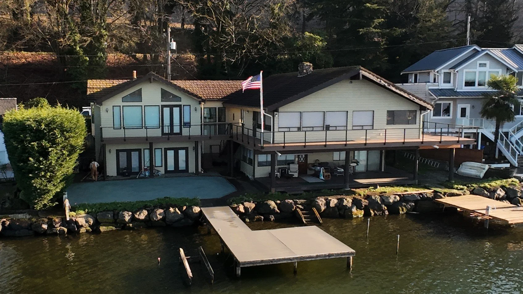 A house by a body of water with a boat dock, deck, and large windows, surrounded by trees and neighboring houses.