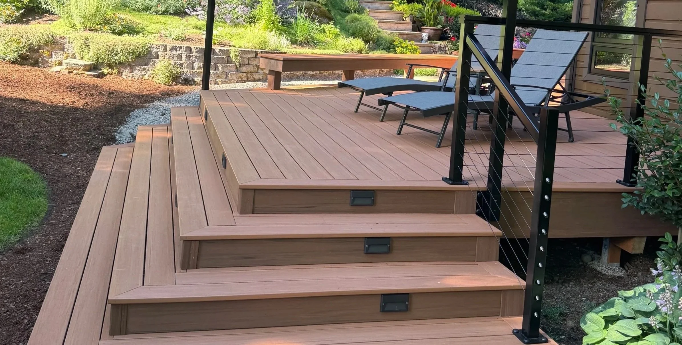 Wooden outdoor deck with steps leading down to garden, featuring a patio table, lounge chairs, surrounding greenery, and a black metal railing.