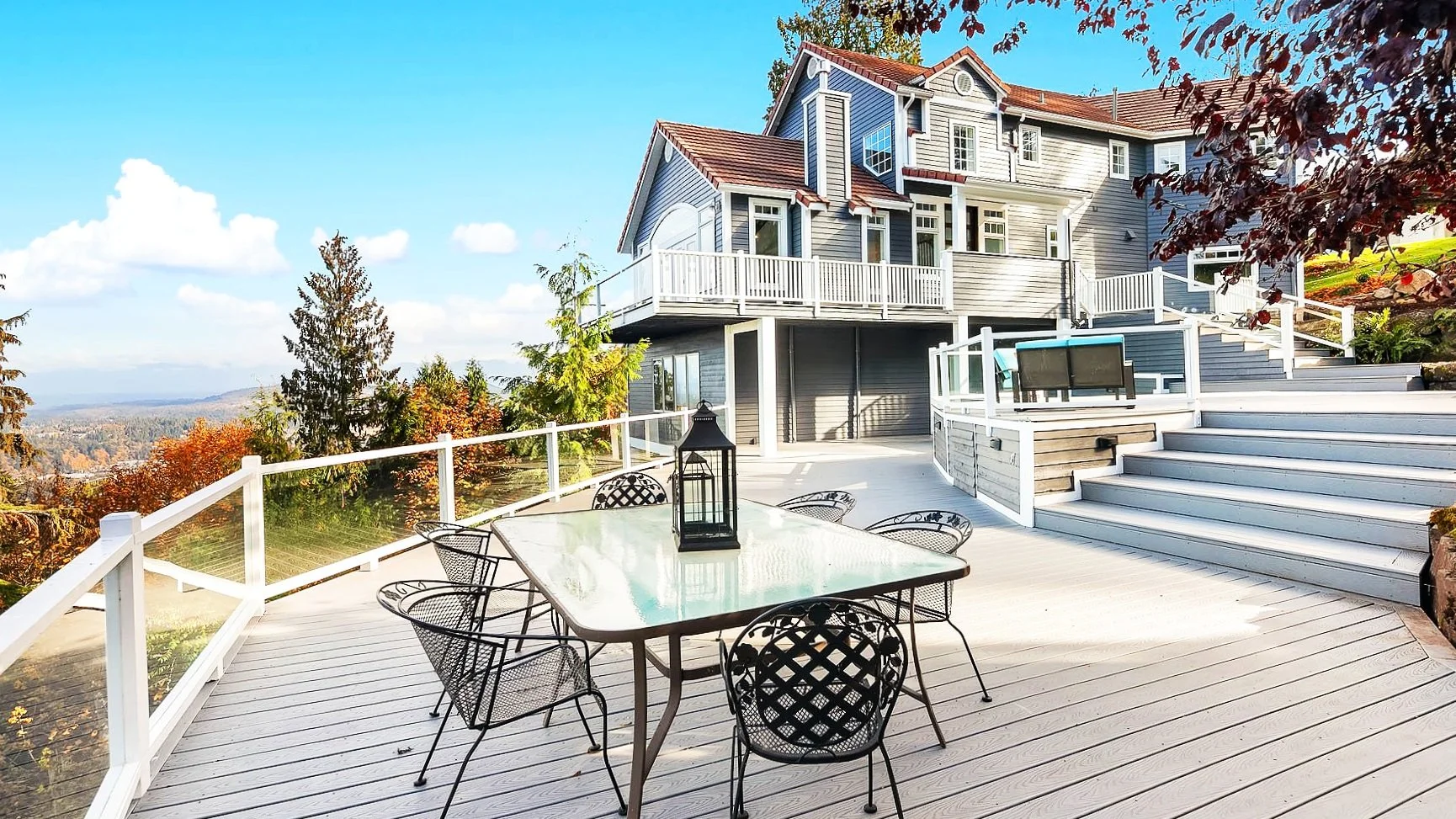 Exterior view of a multi-story house with a spacious wooden deck, black metal outdoor furniture with glass table and lantern centerpiece, stairs leading up to the house, and scenic fall landscape with trees and mountains in the background.