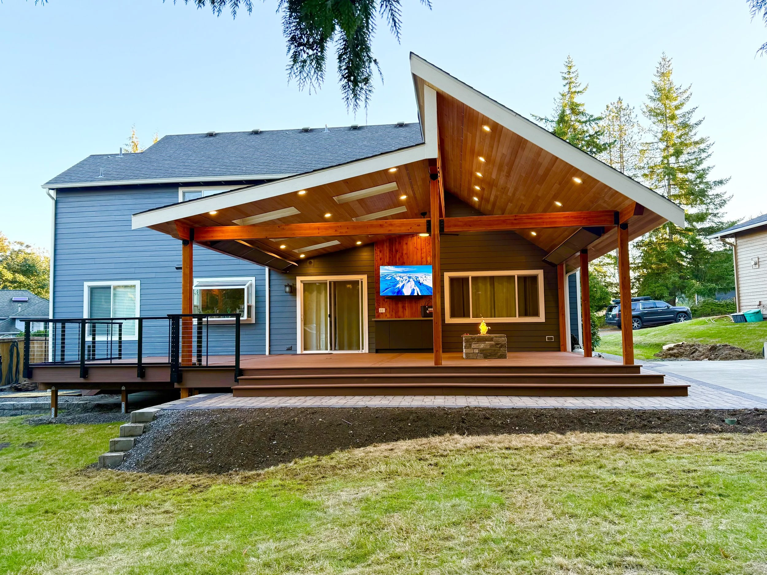 A modern house with a wooden deck and a sloped roof, surrounded by trees, with a flat gravel area and a grassy lawn in the foreground.