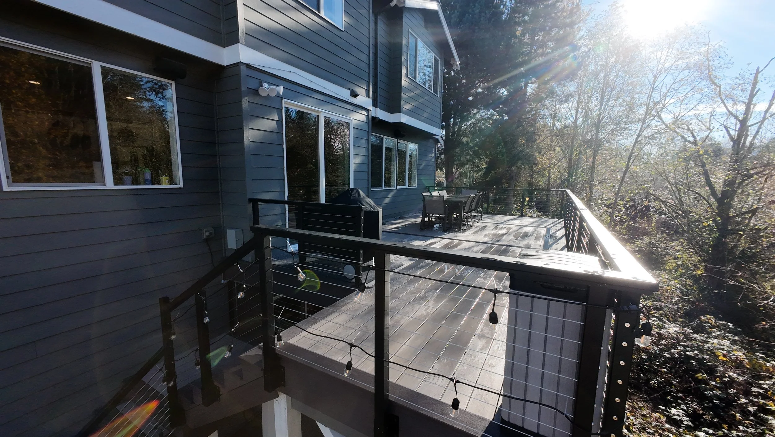Backyard deck with outdoor dining table and chairs, surrounded by trees, with sunlight causing lens flare.