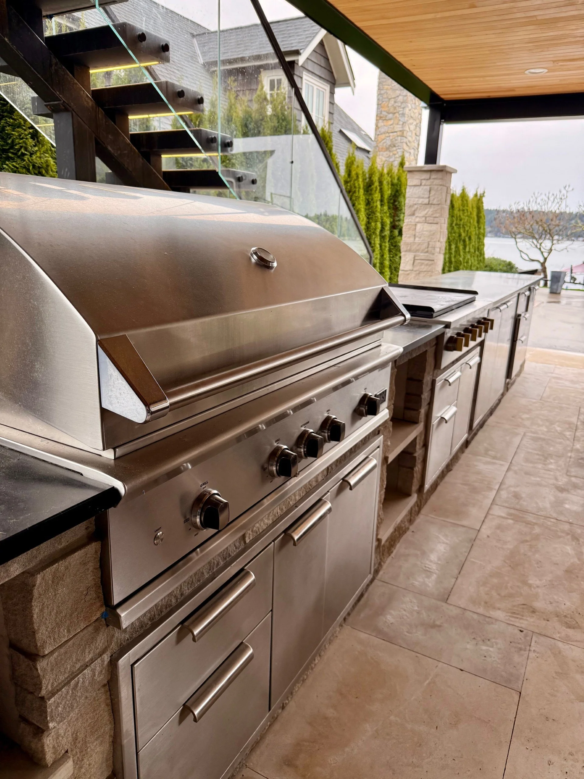 An outdoor kitchen with a stainless steel grill, stone countertop, and cabinetry, with a view of a scenic waterfront in the background.