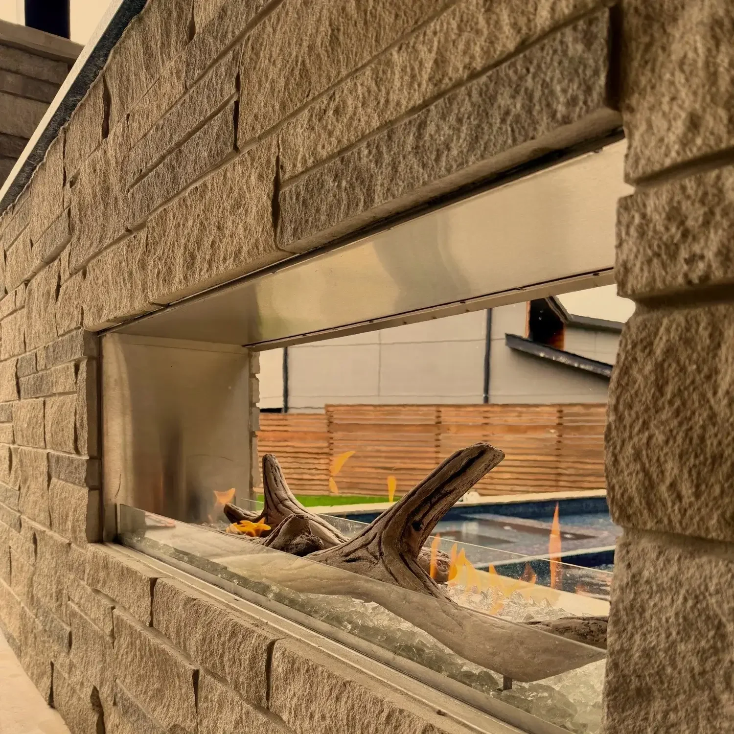 Outdoor linear gas fireplace with stone surround and view of a backyard pool area.