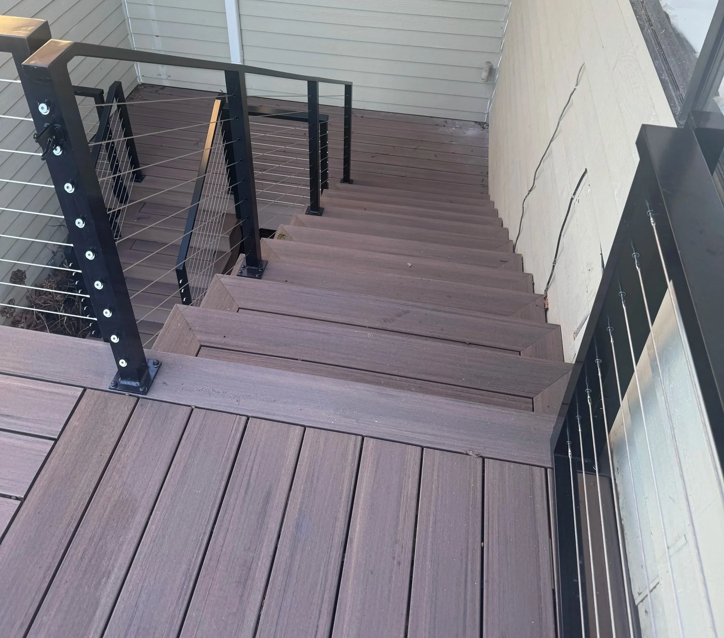 A wooden outdoor staircase with dark wood steps and a black metal railing with thin horizontal wires. The staircase is attached to a light-colored house with vinyl siding and a beige deck. An electrical cord runs along the deck floor, and a glass doo