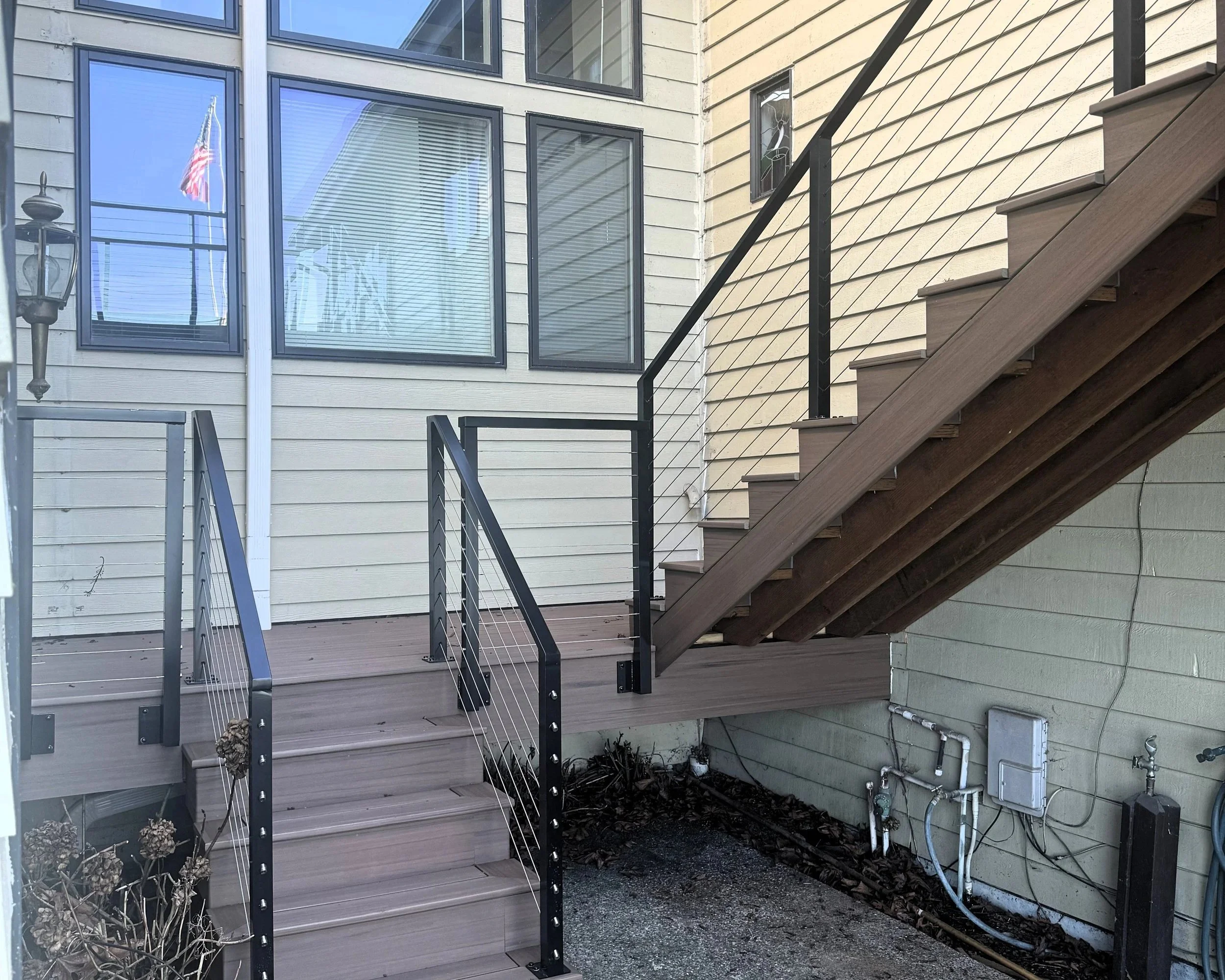 Exterior back porch with gray wood stairs, black handrails, and a small garden area below. The house has beige siding and large windows reflecting the blue sky, with an American flag and a decorative lantern visible through the window.
