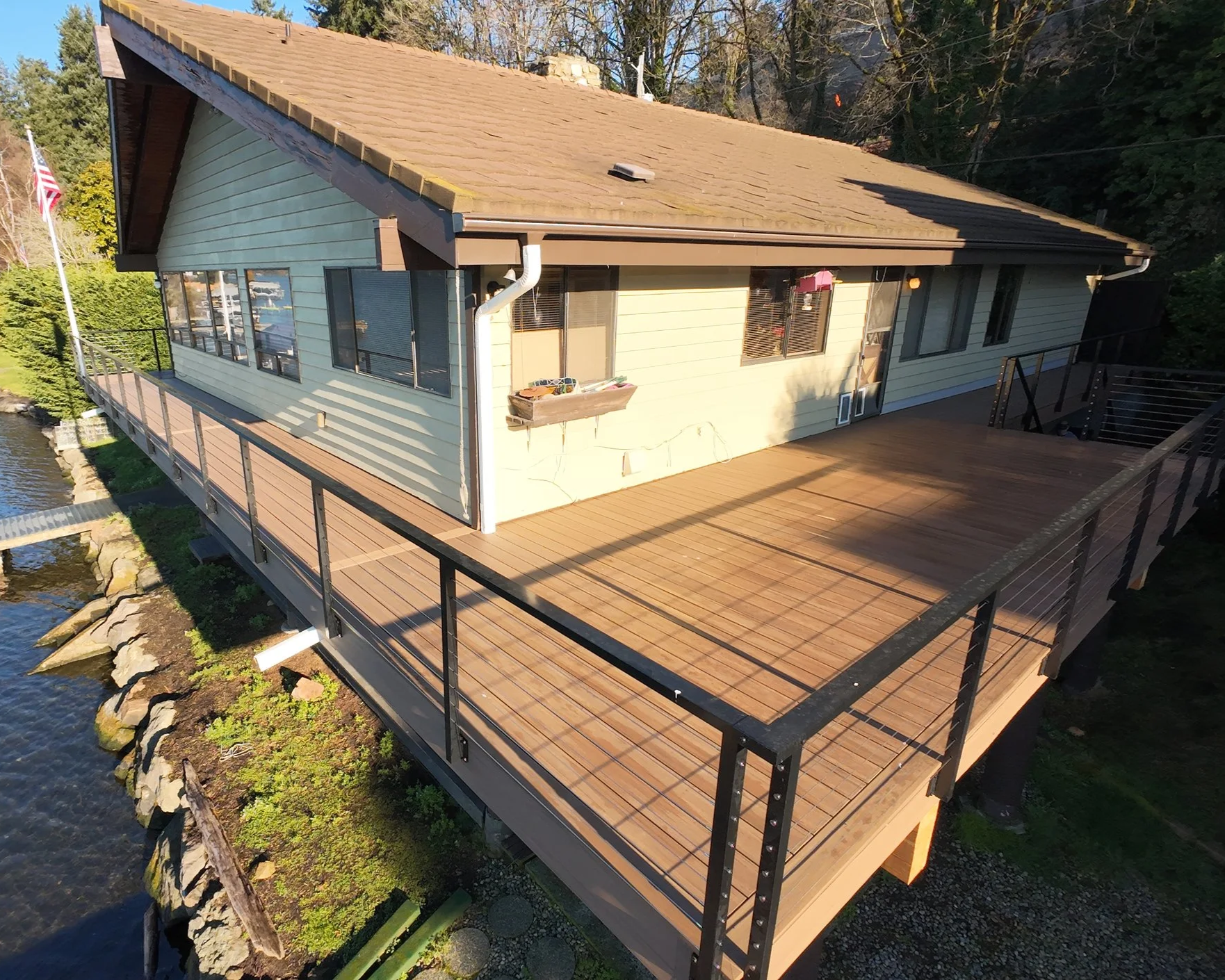 A house with a large wooden deck overlooking a body of water, surrounded by trees and rocks.