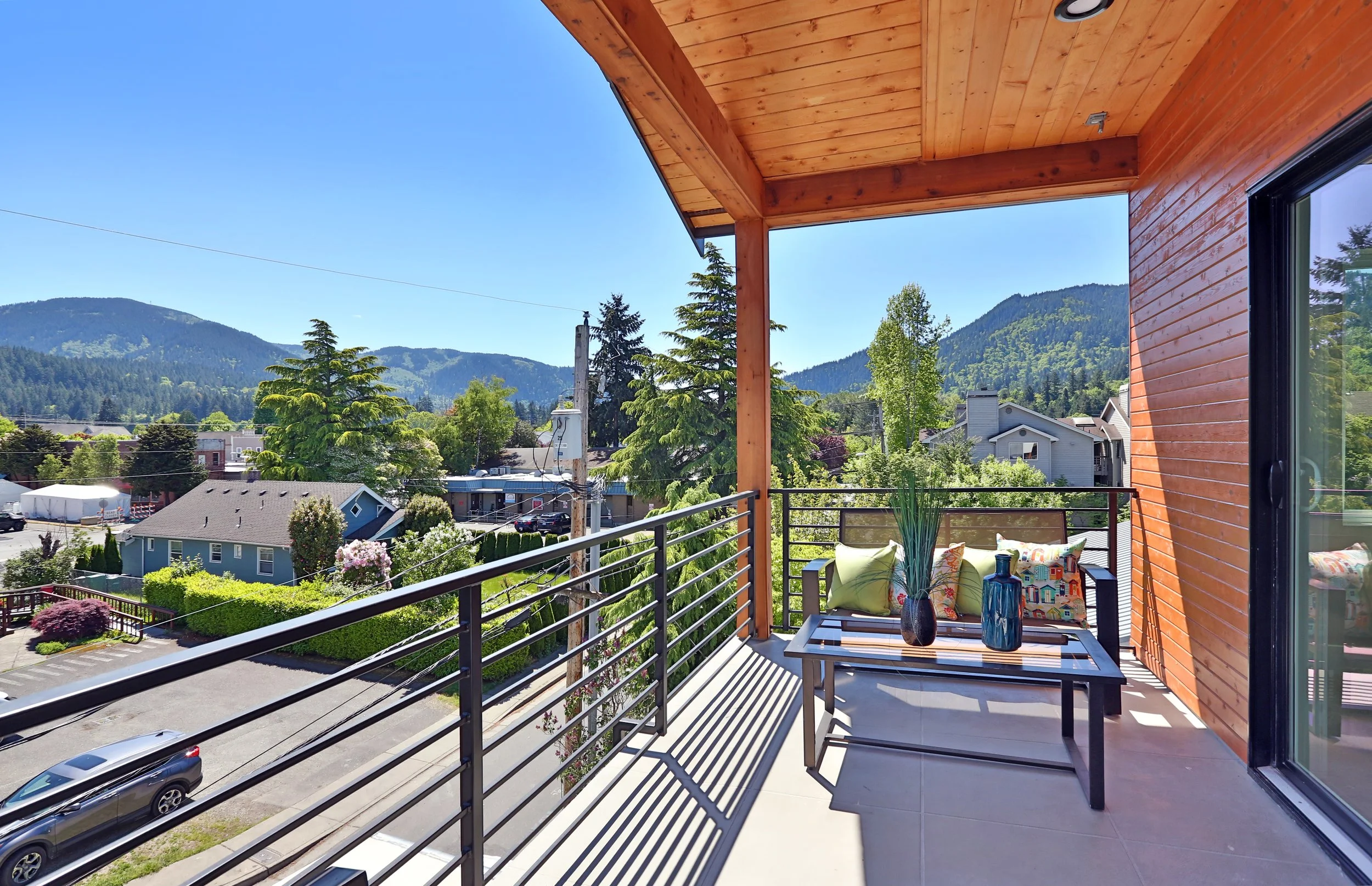 Balcony overlooking a neighborhood with trees and mountains in the distance. The balcony has a black metal railing, outdoor furniture with cushions, and potted plants. The ceiling is wood-paneled.