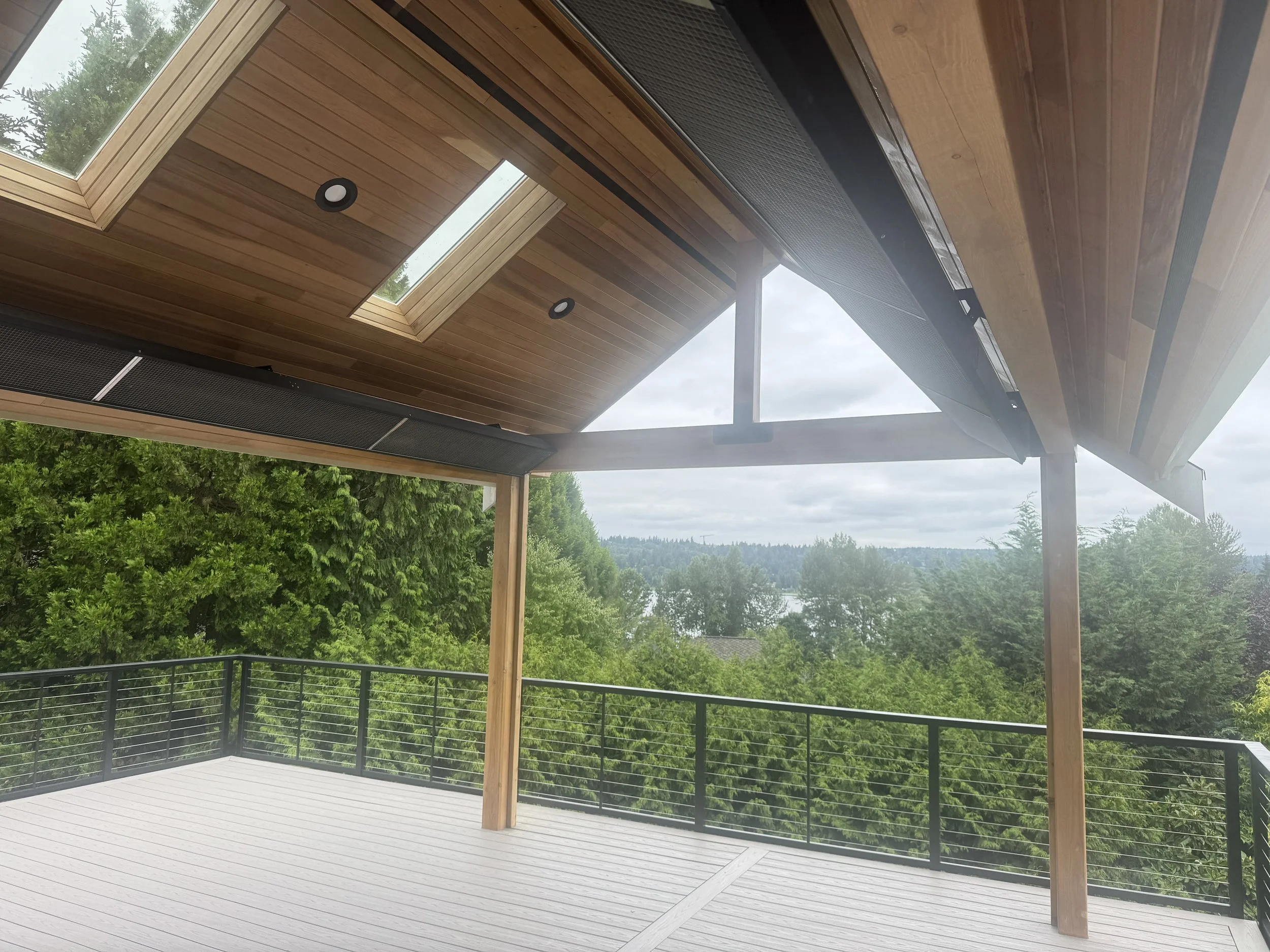 A modern outdoor deck with a wooden ceiling featuring skylights and ceiling lights. The deck has a black metal railing and is surrounded by lush green trees under a cloudy sky.