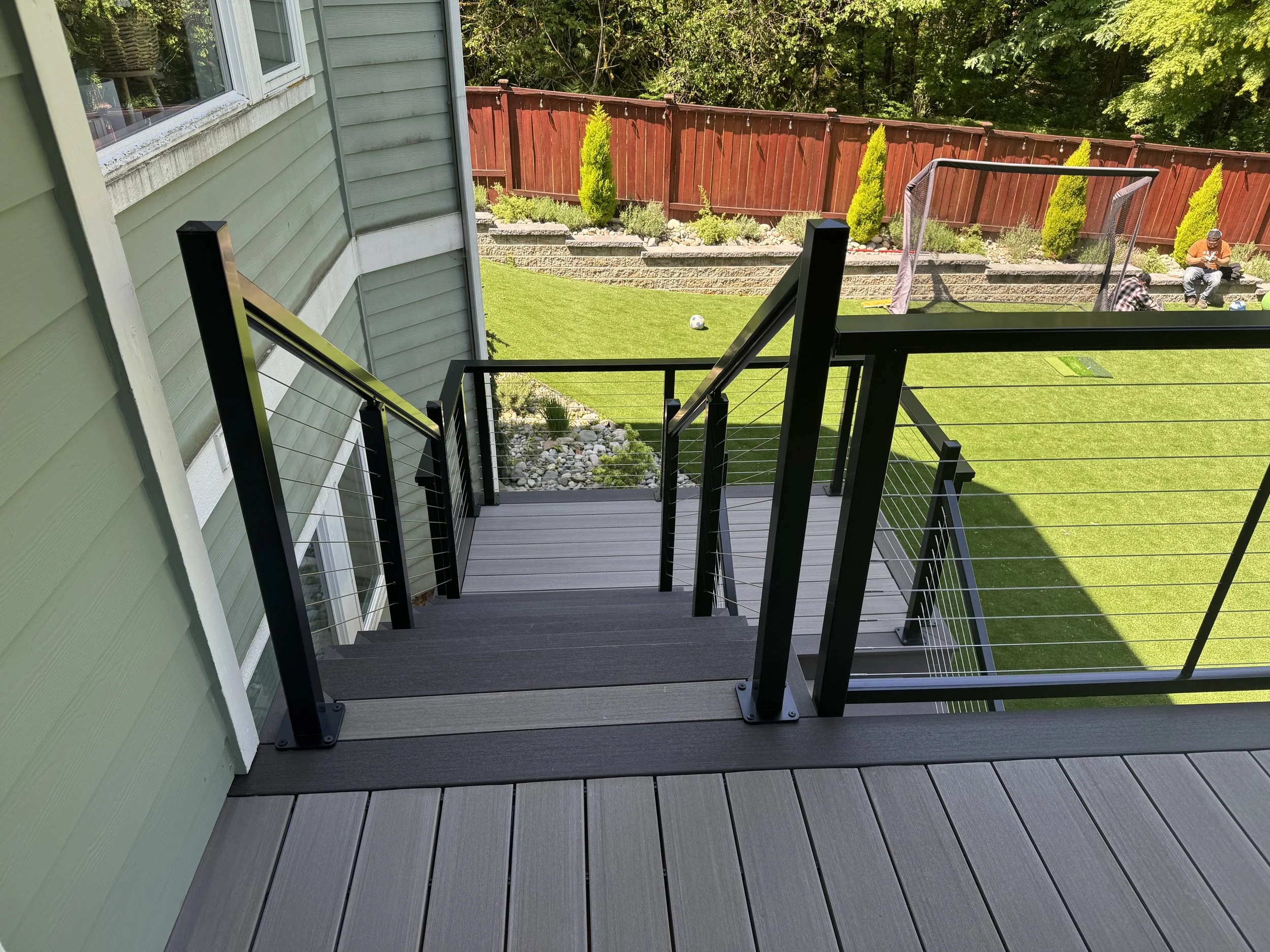View from a balcony looking out onto a backyard with a grass lawn, deck chairs, and a red wooden fence with plants and trees behind it.