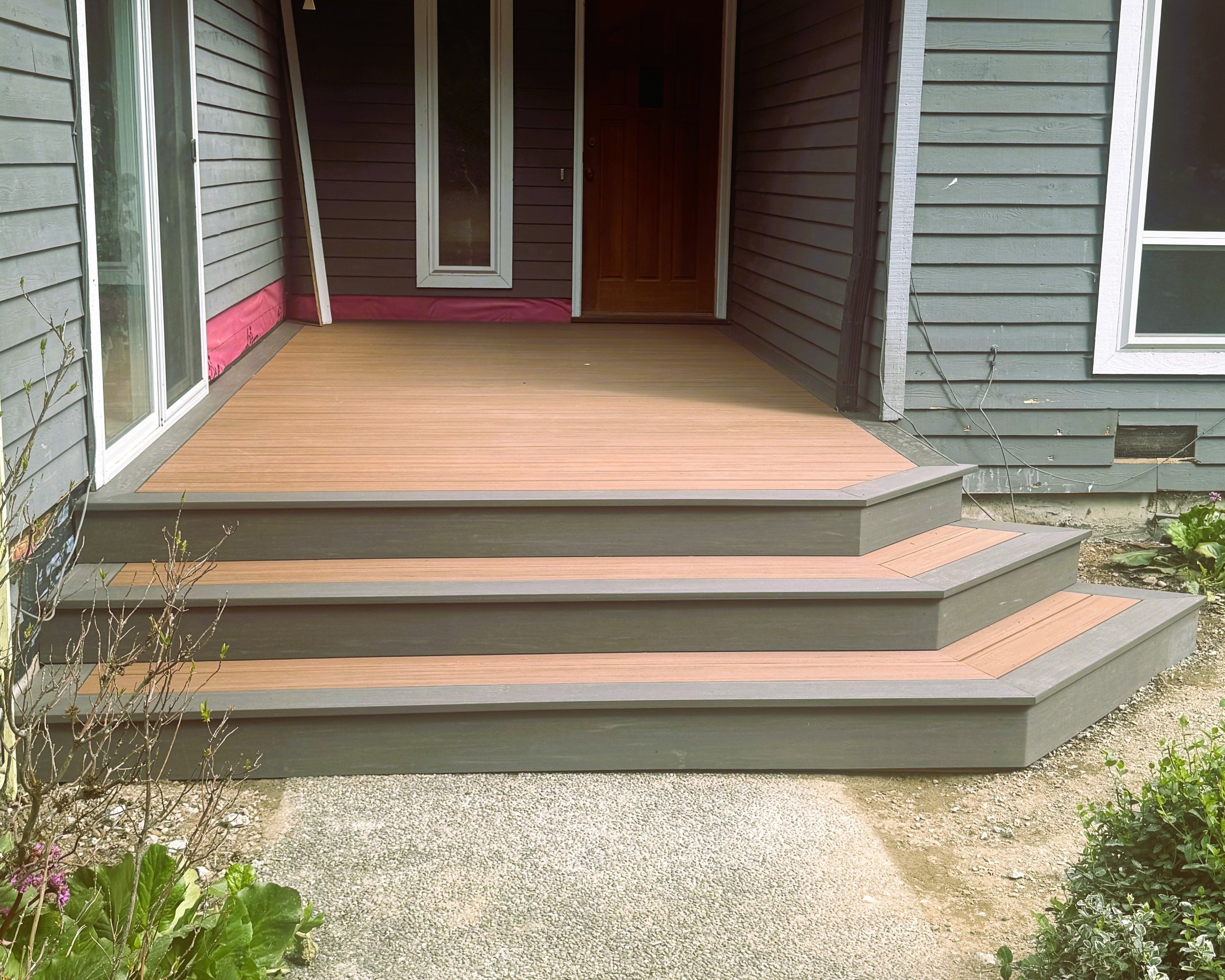 An outdoor wooden deck attached to a house with grey siding, a sliding glass door, a front door, and some small plants and flowers nearby.