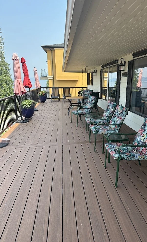 A spacious balcony with wooden decking, four floral-patterned chairs, a small table, potted plants, and closed red and pink umbrellas, overlooking a residential area with other buildings.