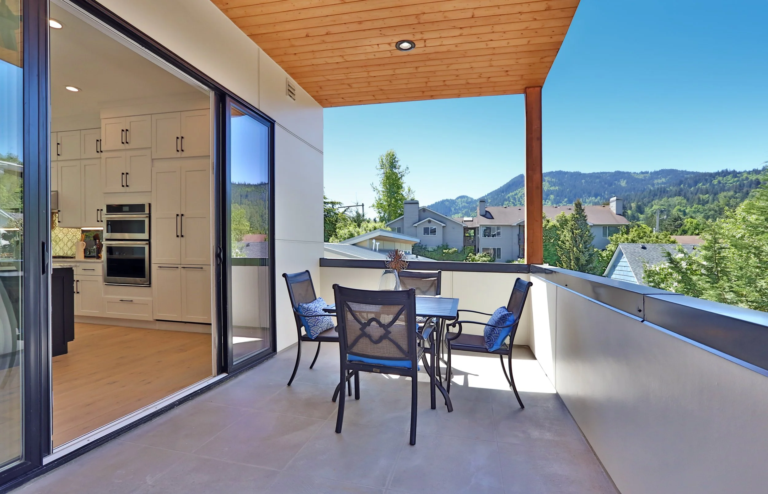 Balcony with outdoor dining table and chairs overlooking a suburban neighborhood with houses, trees, and mountains in the background, under a clear blue sky.