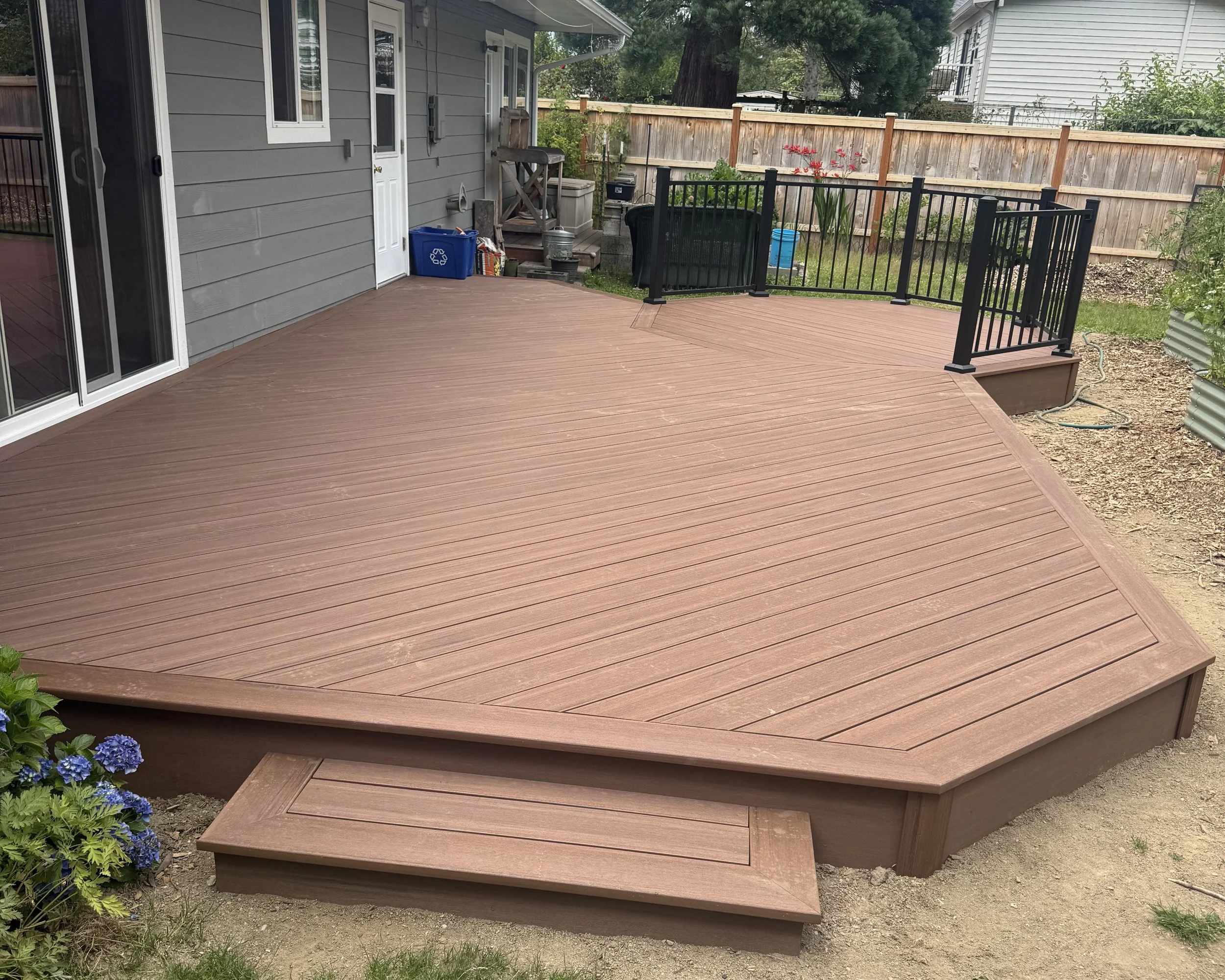 A backyard with a wooden deck attached to a house with gray siding. The deck has a small set of stairs and a black safety gate. There are some plants and flowers along the fence, with a large tree in the background. Some gardening tools and items are