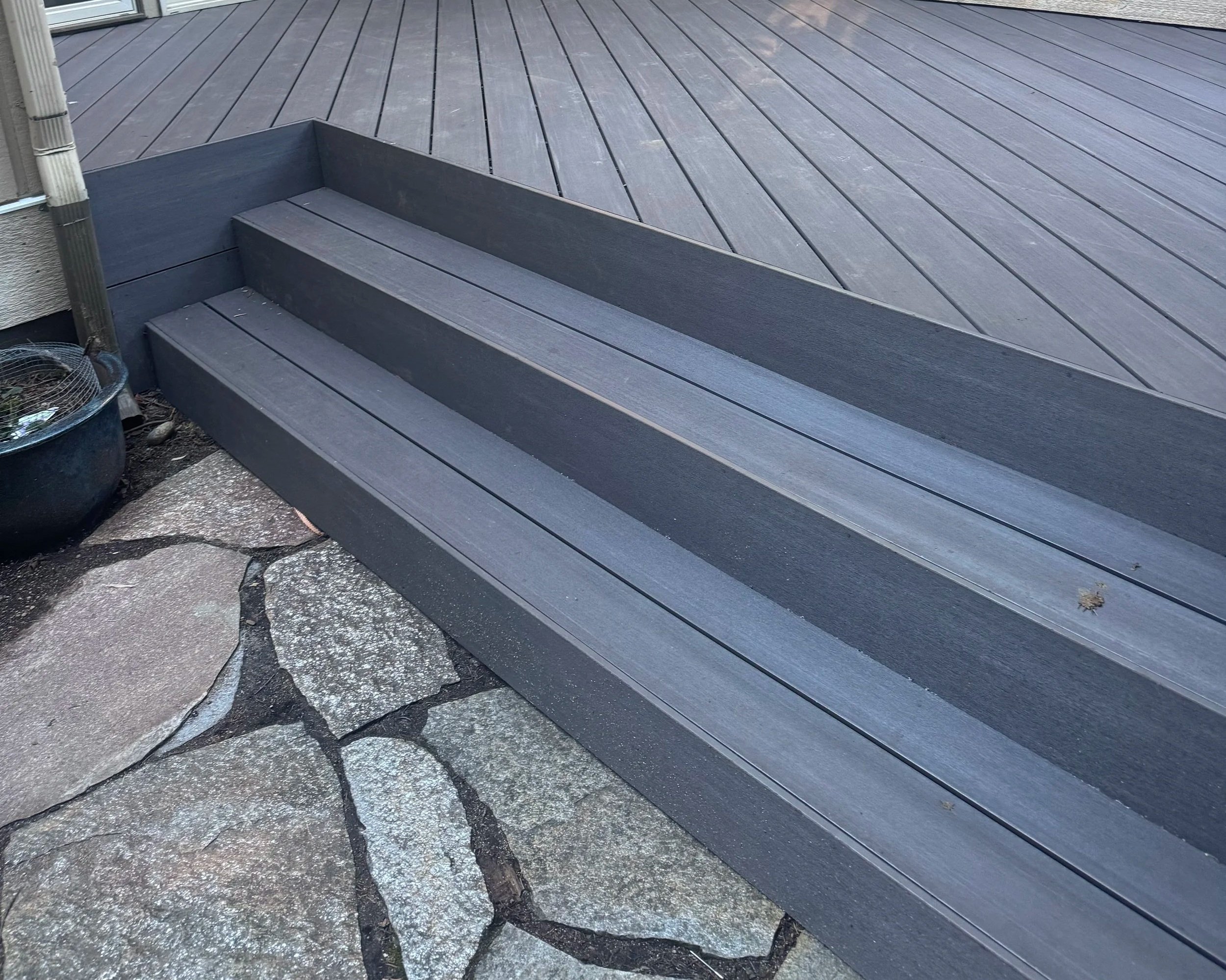 A wooden deck with stairs and a section of concrete pavement with cracked and uneven surface, near a building with a window.