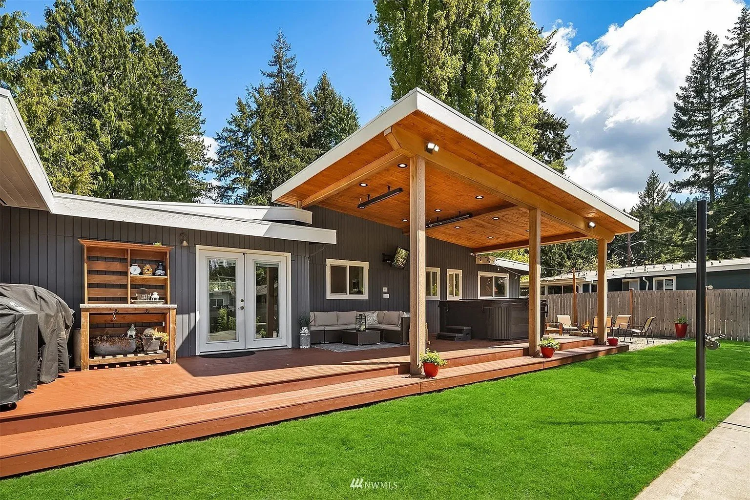 Backyard with a wooden deck, covered seating area, hot tub, outdoor furniture, green lawn, and tall trees in the background.