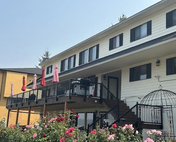 Backyard view of a two-story house with a large balcony, pink and red roses in the garden, and a metal birdcage near the stairs.