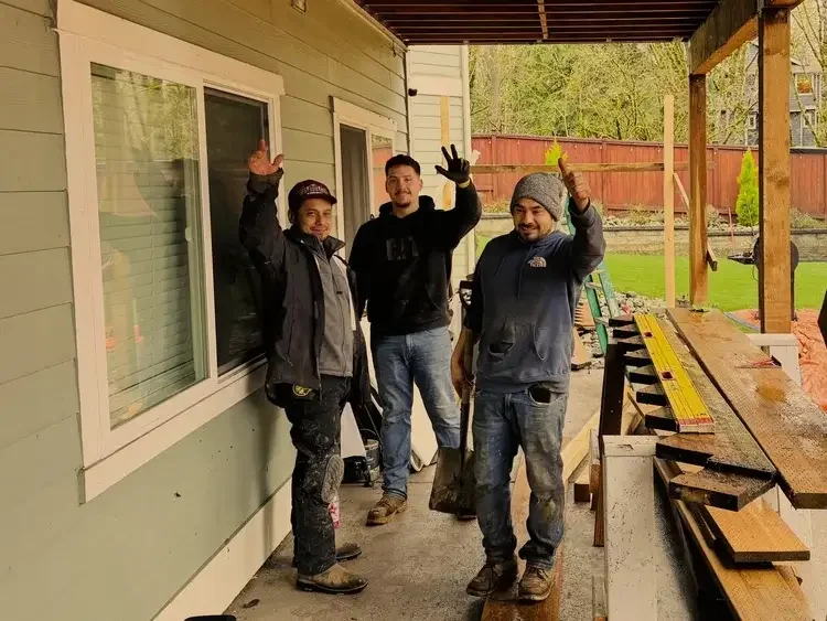 Three construction workers standing on a porch next to a house, giving a thumbs-up gesture. They are surrounded by construction materials and tools, with a partially constructed structure in the background.