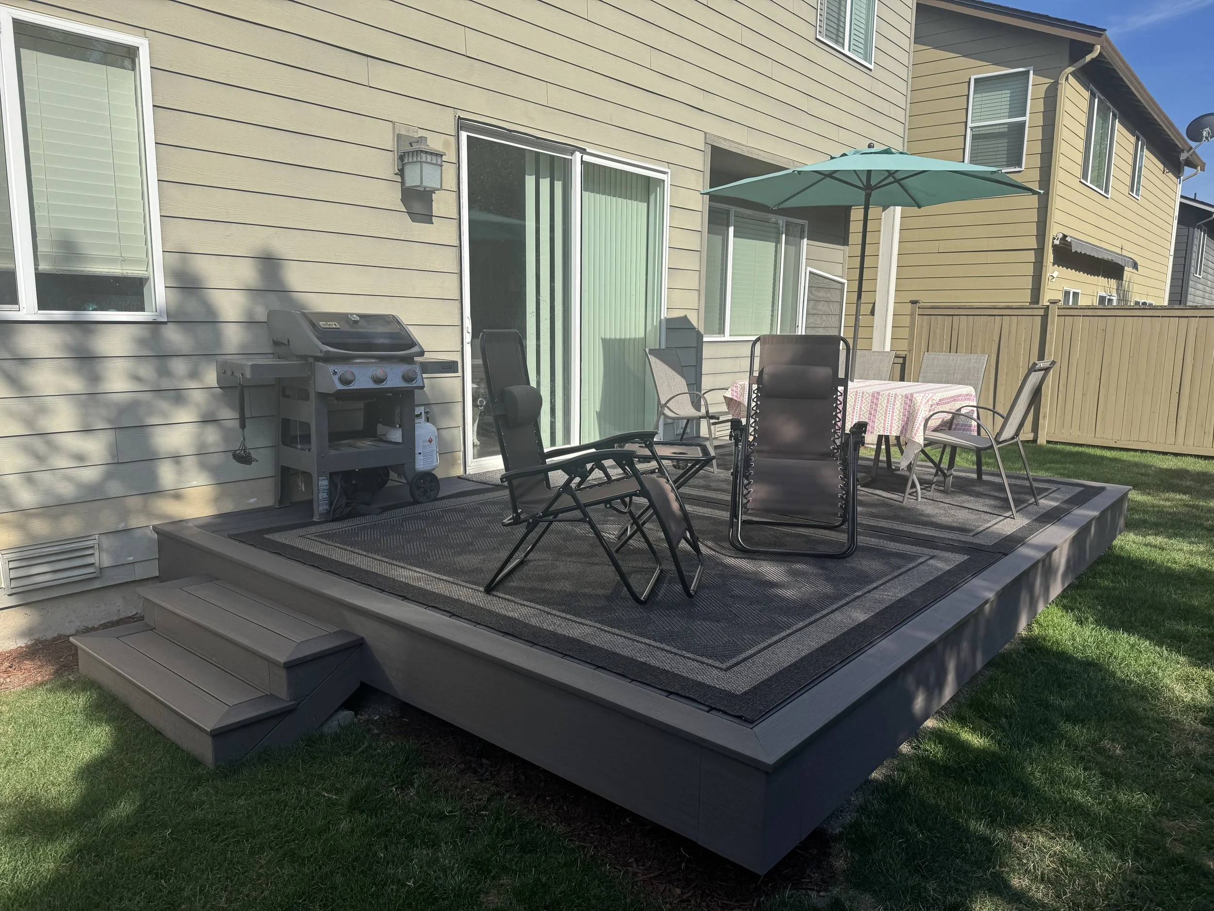 A backyard patio with outdoor seating, a table with a tablecloth, an umbrella, a grill, and a sliding glass door leading into a house.