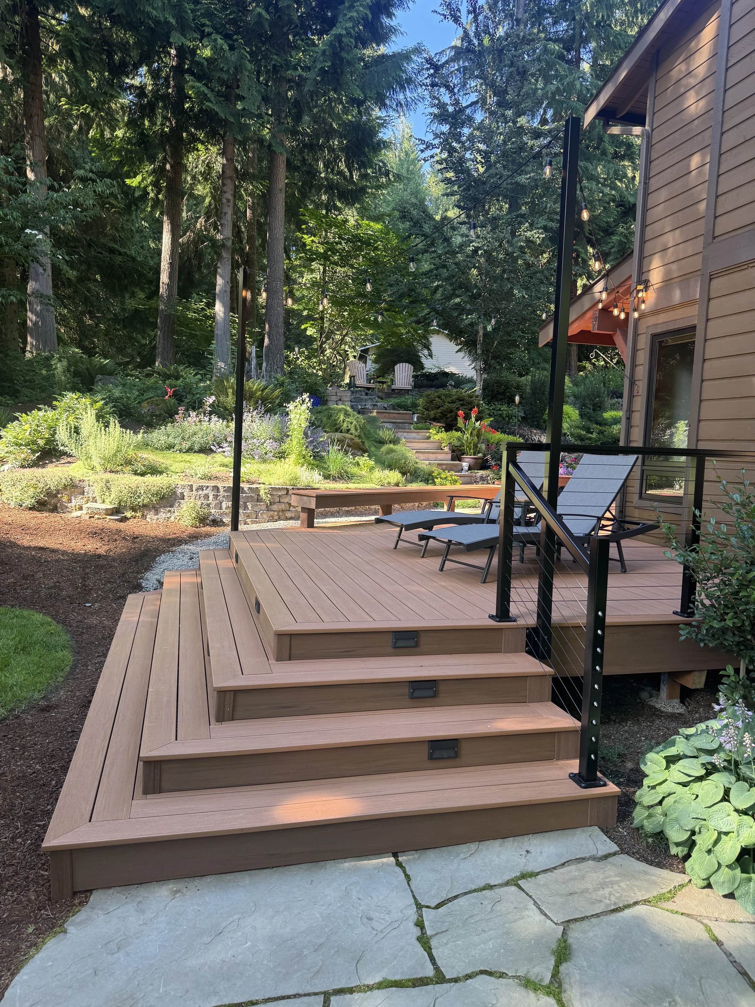 A wooden deck with steps leading down to a stone patio, surrounded by lush greenery and trees, with outdoor chairs and string lights hanging above.