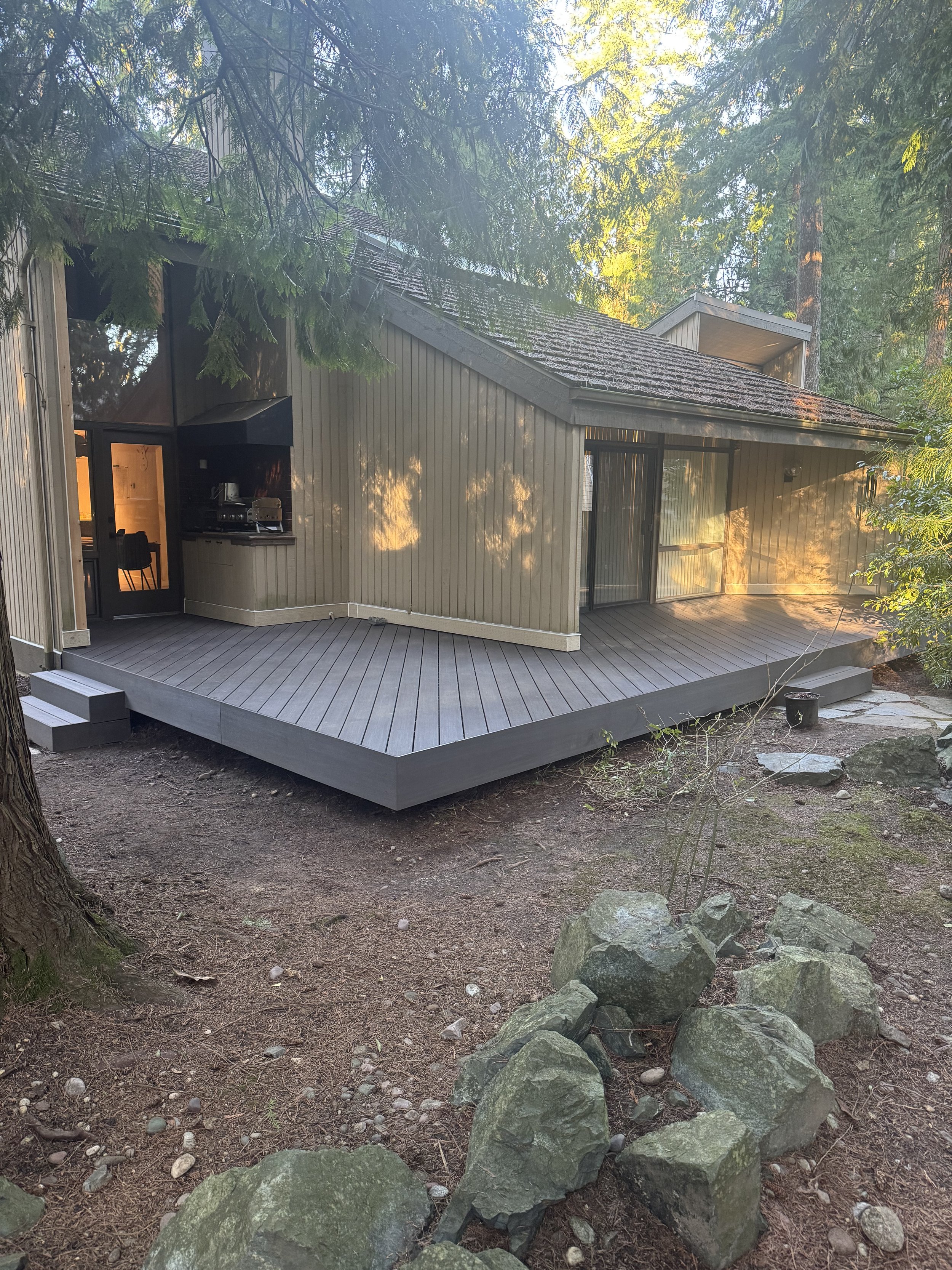 A modern house with wood siding and large glass doors, surrounded by tall trees and rocks, located on a wooden deck in a forest.