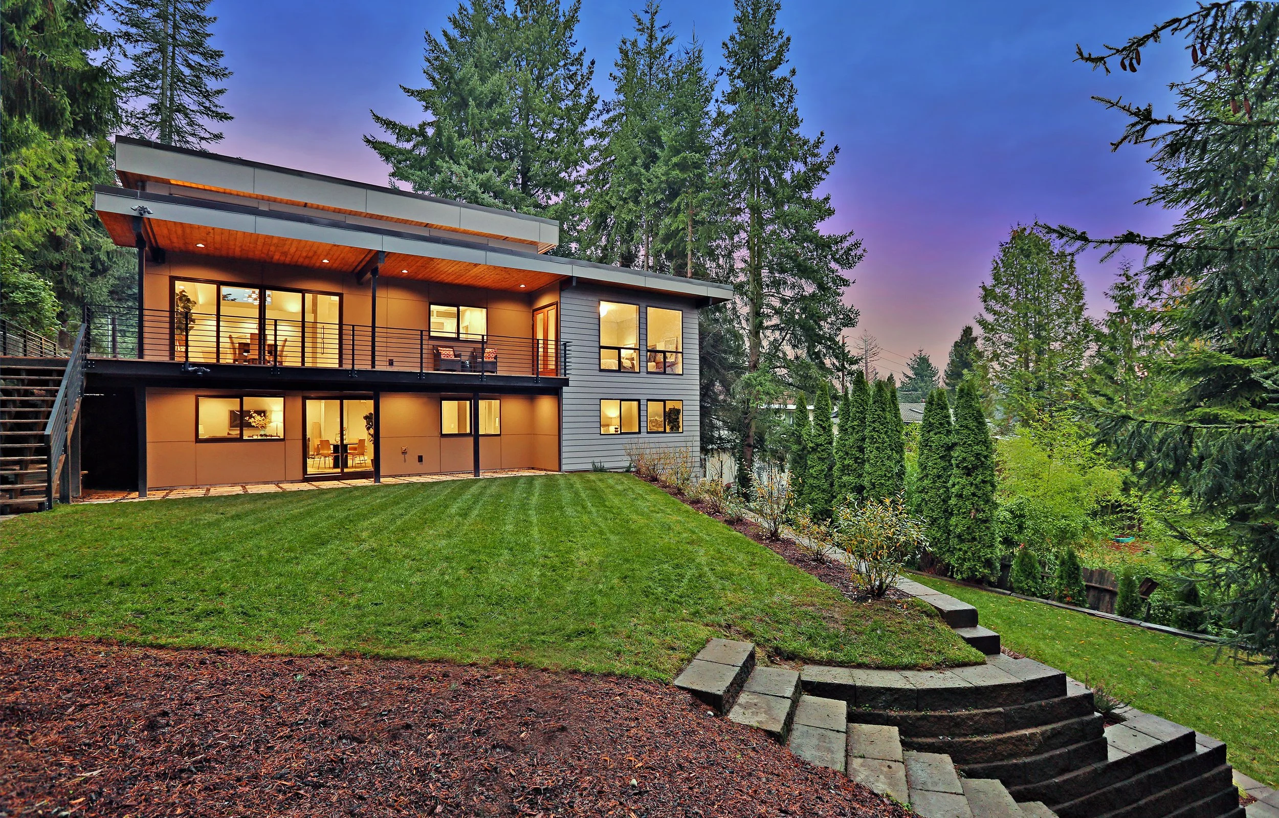 Modern house with large windows and a balcony, surrounded by tall evergreen trees and a well-maintained grassy lawn at dusk.