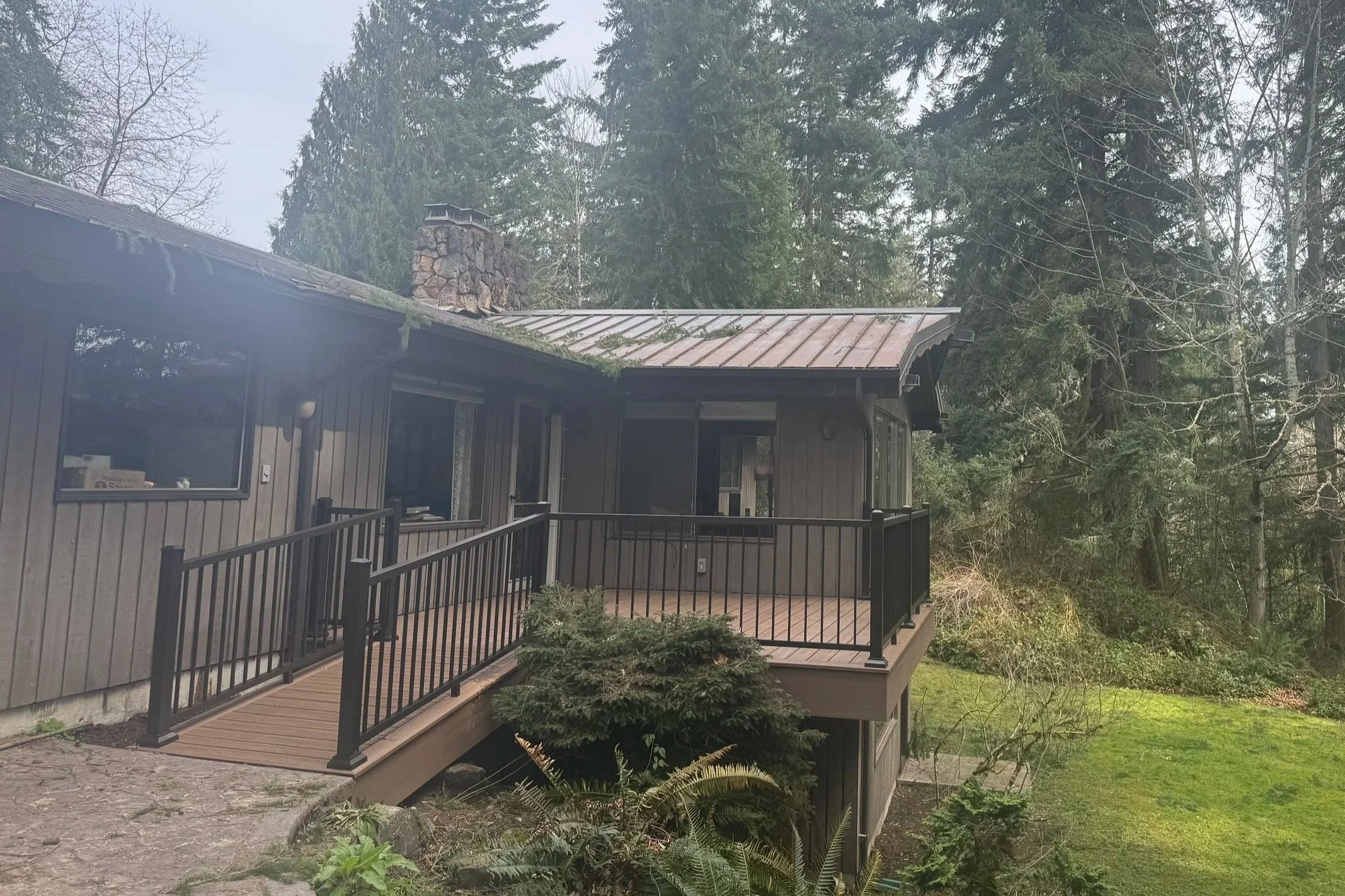 A house with dark brown siding and a metal roof, with a deck and black railing, surrounded by trees and greenery in a forested area.