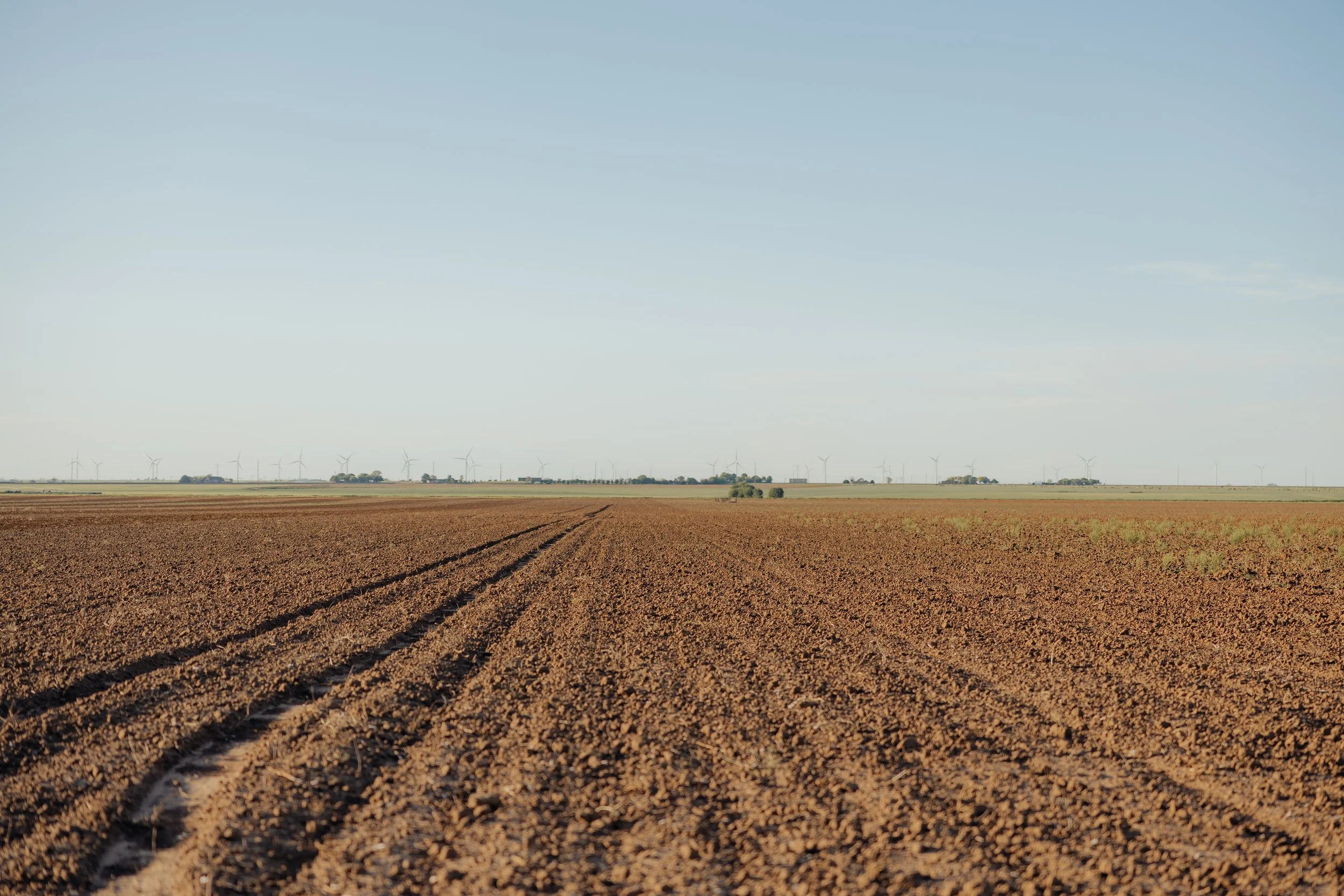 A barren farmland field with soil, and wind turbines in the distance against a light blue sky.