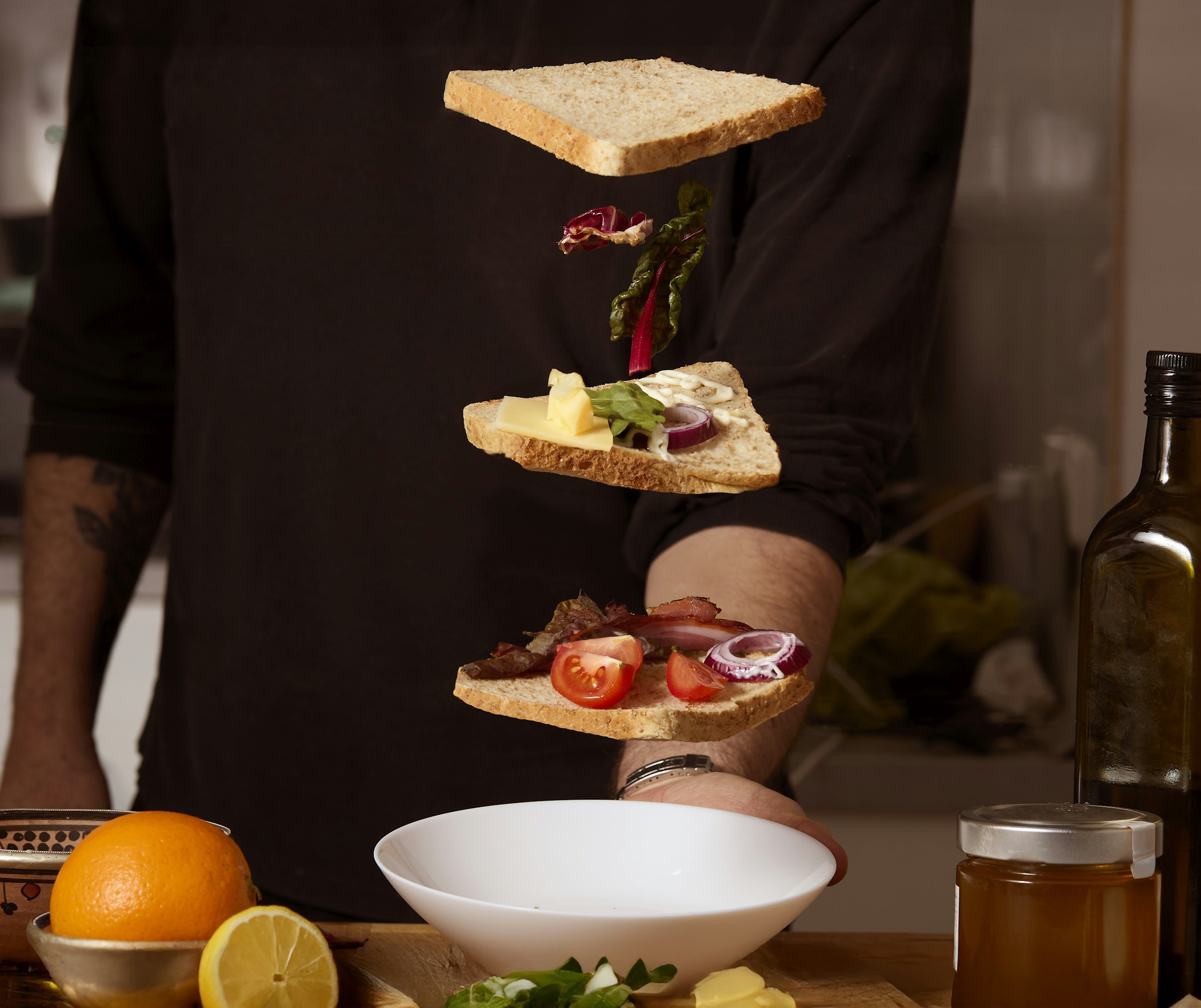 A person stacking three slices of bread with various ingredients. The sandwiches include vegetables, cheese, and meats, with fresh ingredients and a bowl and condiments nearby.