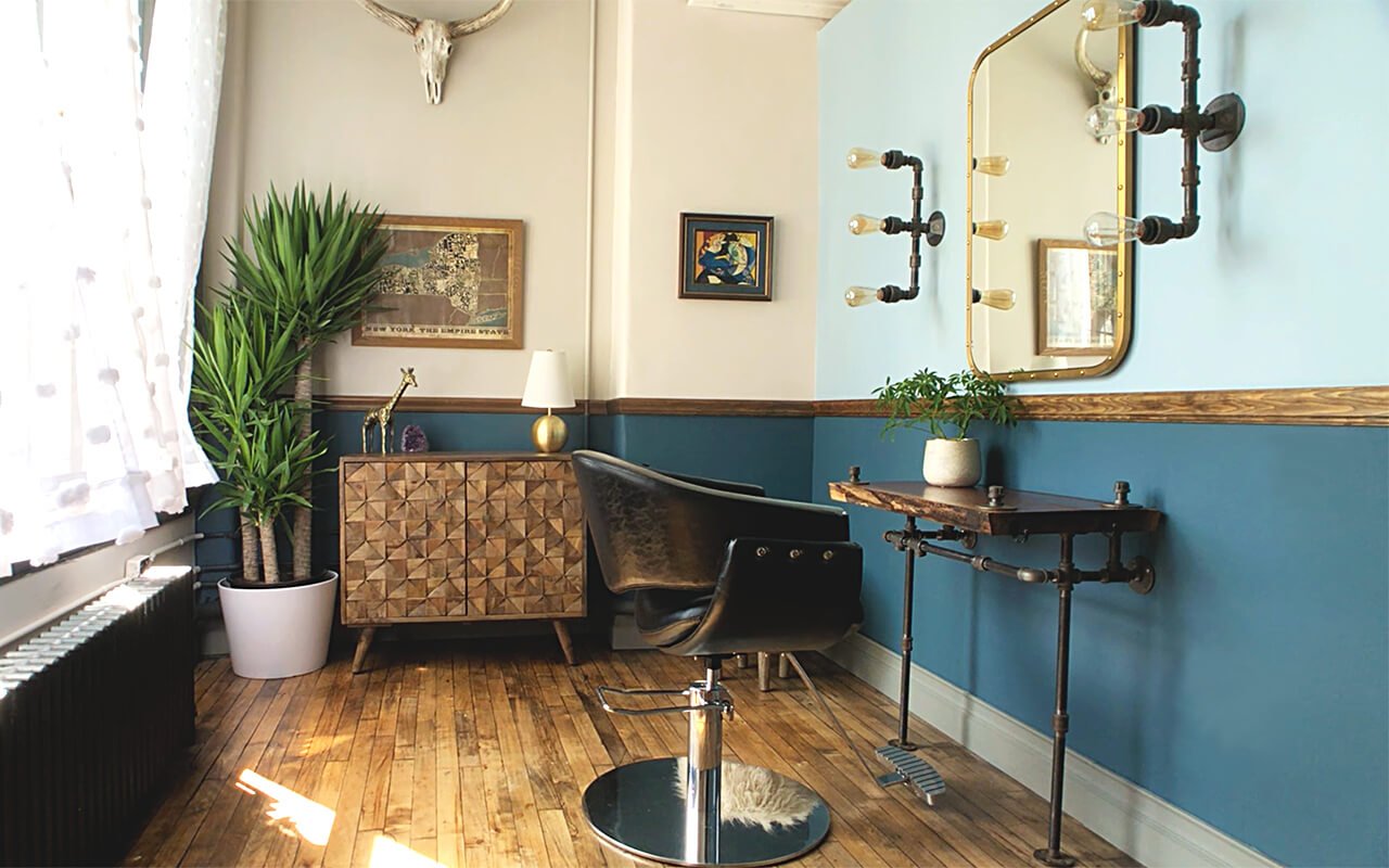 Stylish salon suite with wooden flooring, industrial pipe lighting, vintage mirror, potted plants, decorative wall art, and a leather styling chair.