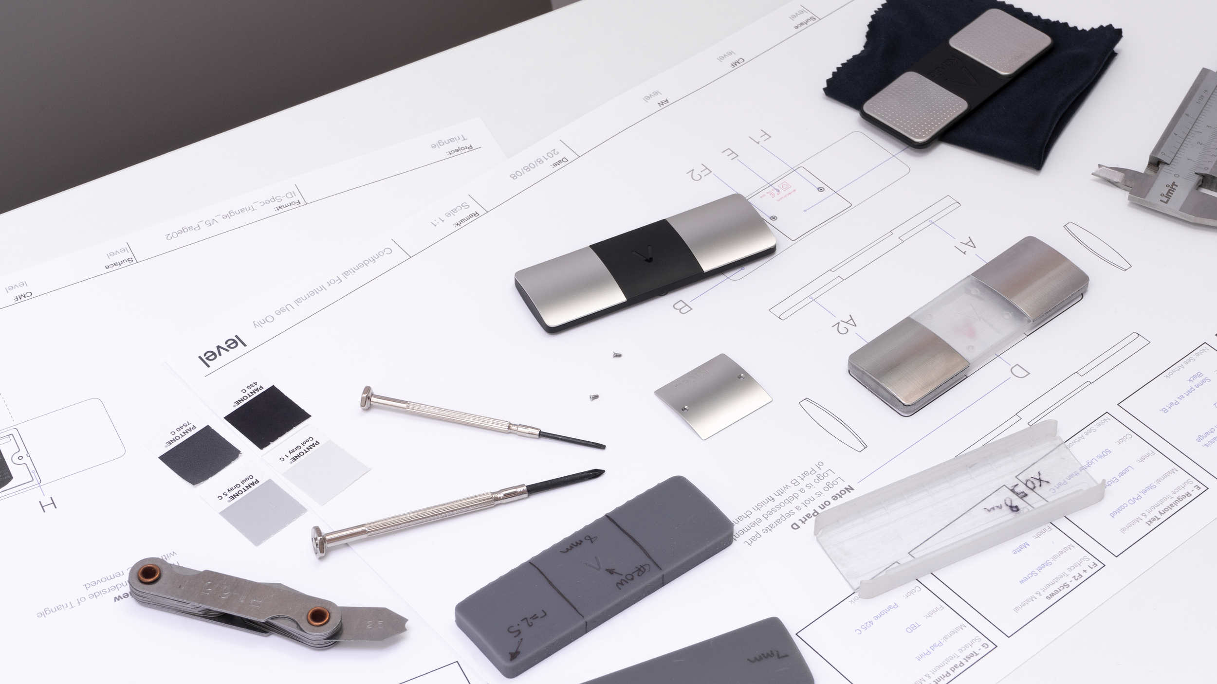 Tools and samples placed on an engineering blueprint, including a caliper, screwdrivers, metal pieces, and color swatches.
