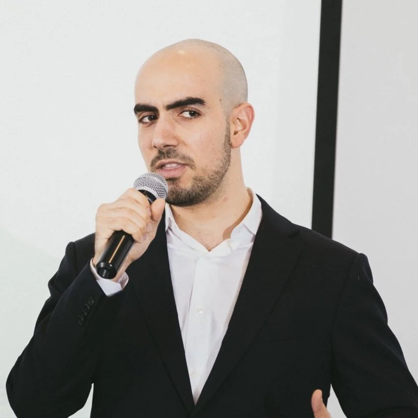 A man with a bald head and beard wearing a black suit and white shirt, holding a microphone and speaking.