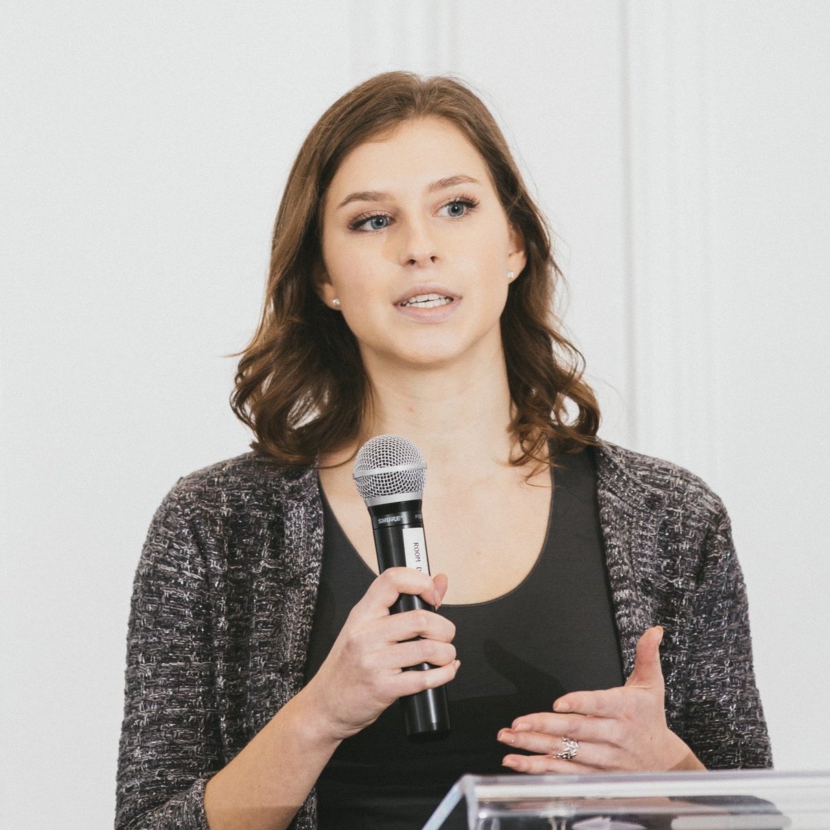 A woman with brown, shoulder-length hair wearing a dark top and a patterned blazer, holding a microphone and speaking.