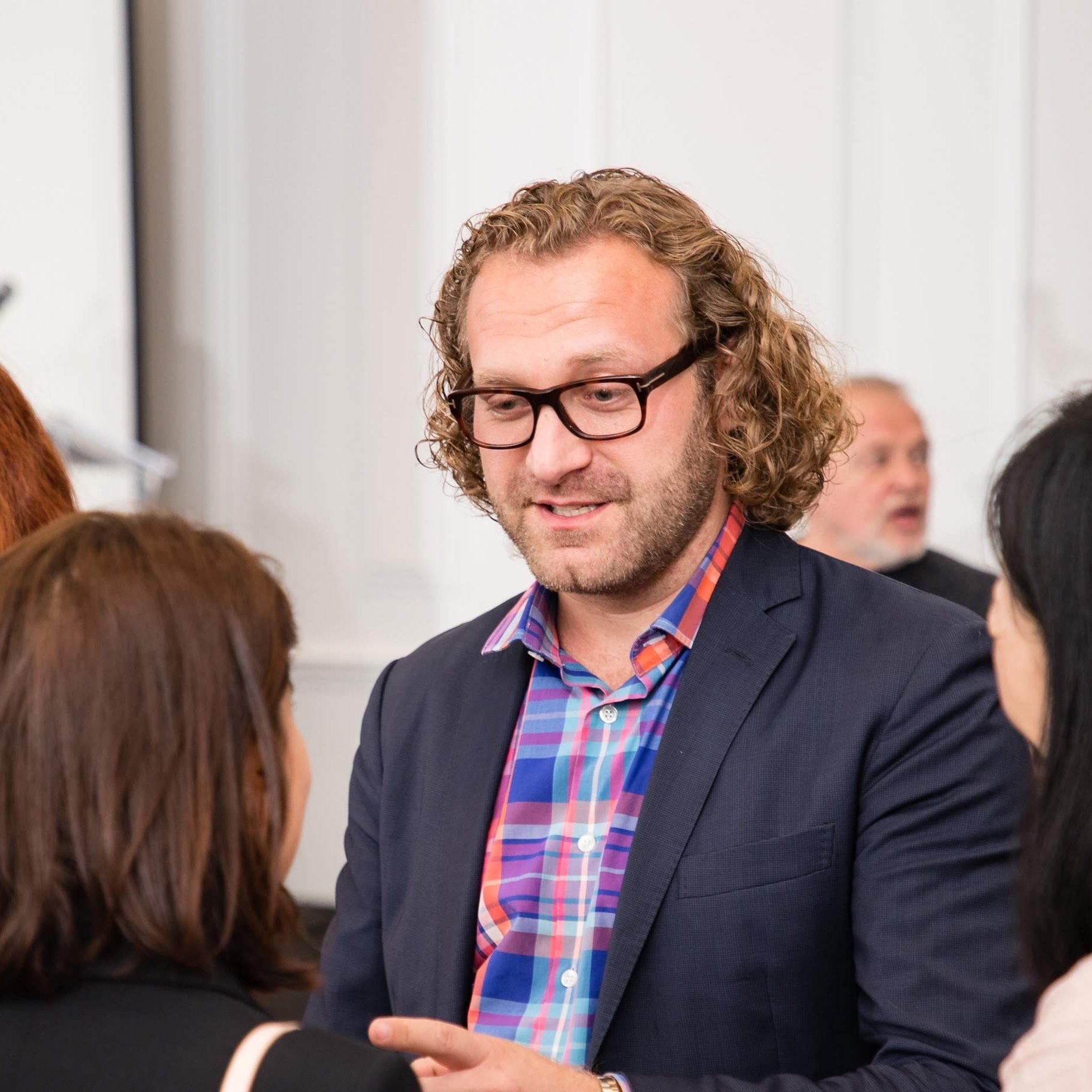 Man with curly hair, glasses, and a plaid shirt talks to women at a social event.