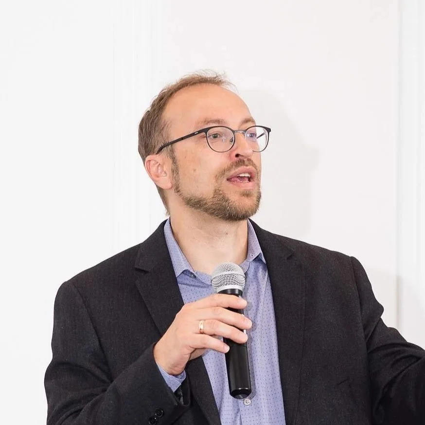 Man in black blazer speaking into a microphone at a presentation or meeting.