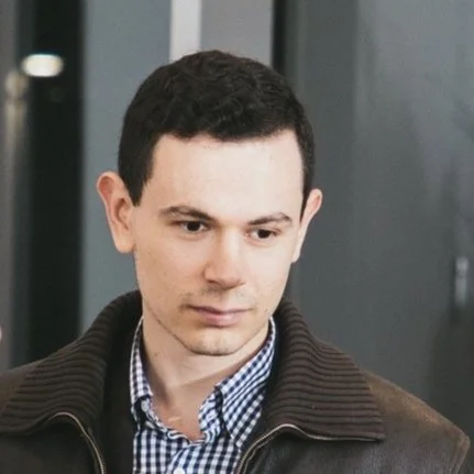 A young man with short dark hair, wearing a checkered shirt and a dark jacket, looking downward in an indoor setting.
