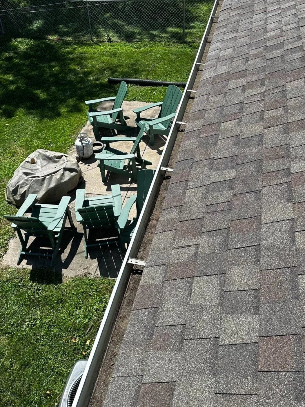 View from the roof showing a backyard with green lawn, a fenced area, four green wooden chairs, a covered grill, and a propane tank.