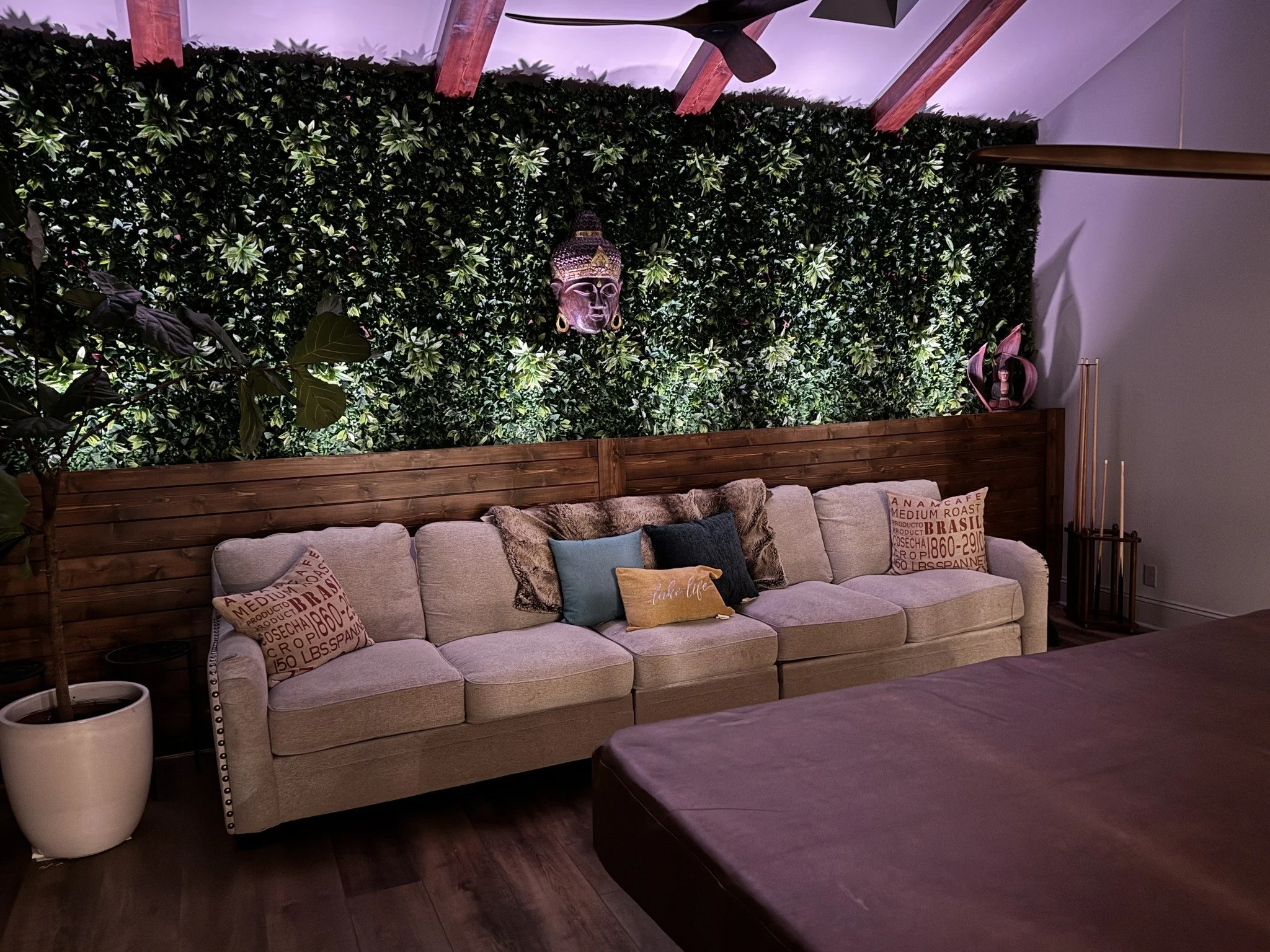 Living room with a beige sofa decorated with various pillows, a green leafy wall with a Buddha face mask, and wooden ceiling beams.