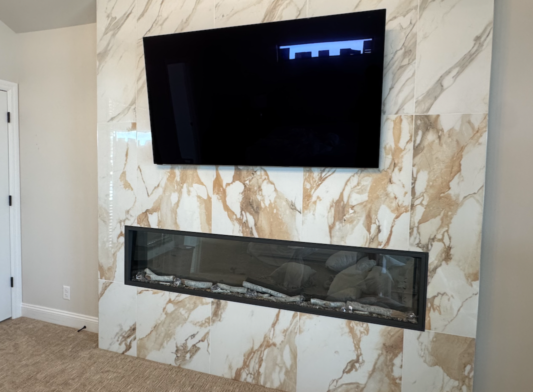 A flat-screen TV mounted above a modern glass fireplace with decorative rocks, on a wall with polished marble tiles.